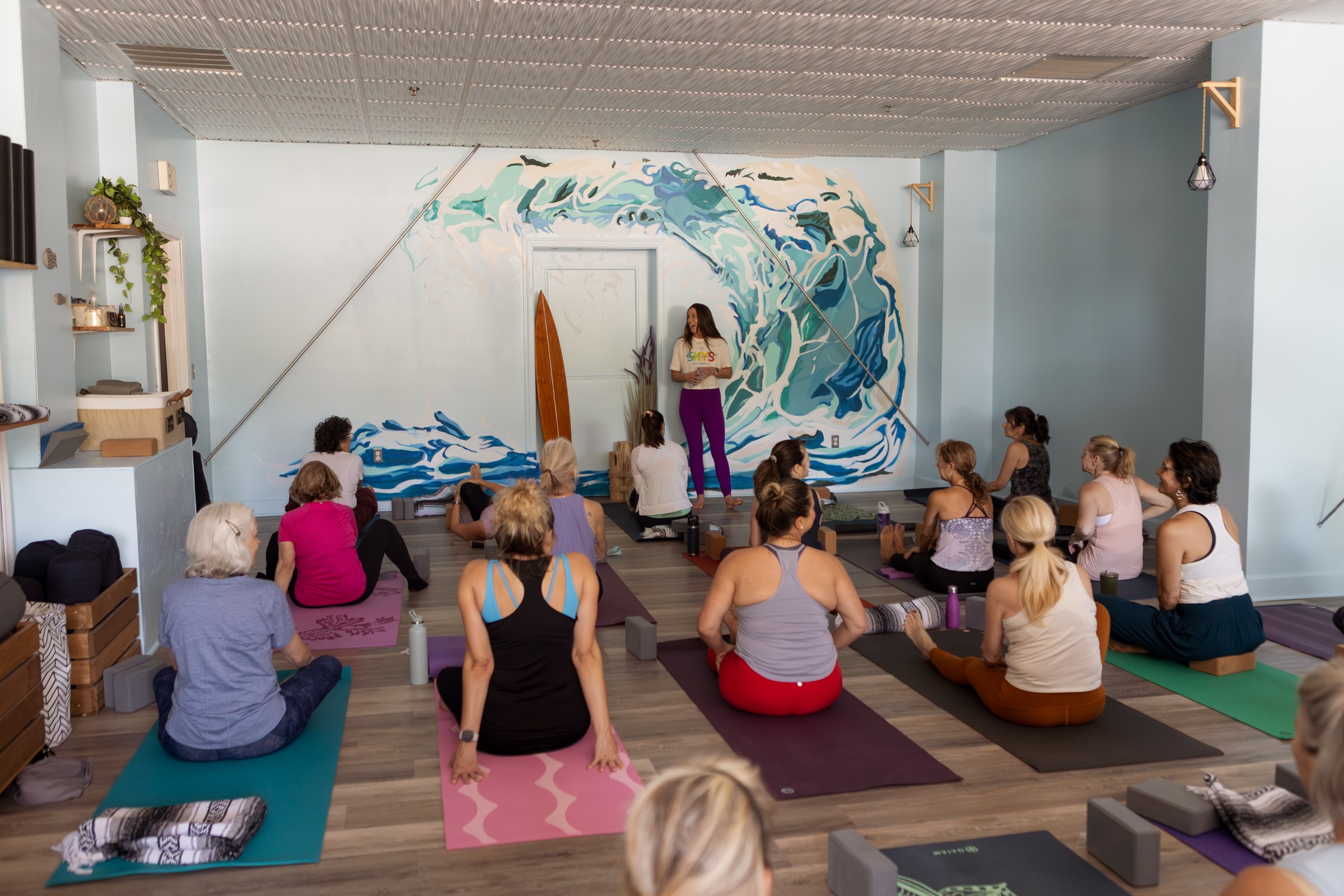 Group of people participating in a yoga or meditation class in a room with ocean-themed mural, sitting on mats facing instructor.