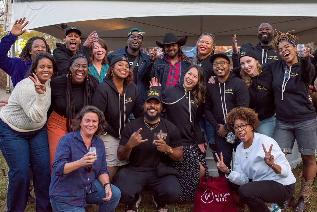 Group of diverse people smiling, posing outdoors, some making peace signs, in casual clothing, in front of a building.