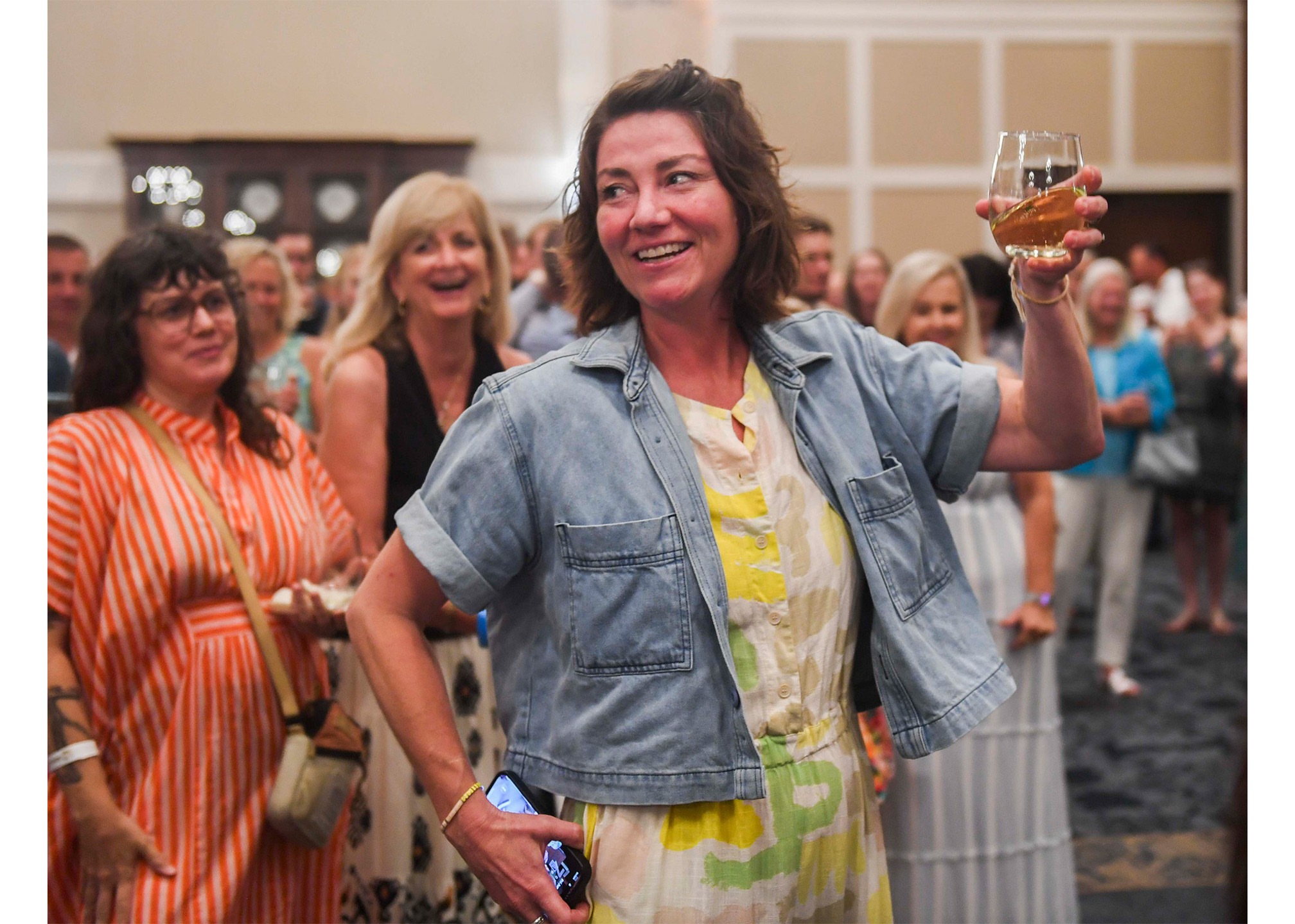 Woman smiling and holding a glass of drink at a social gathering, surrounded by other people indoors.