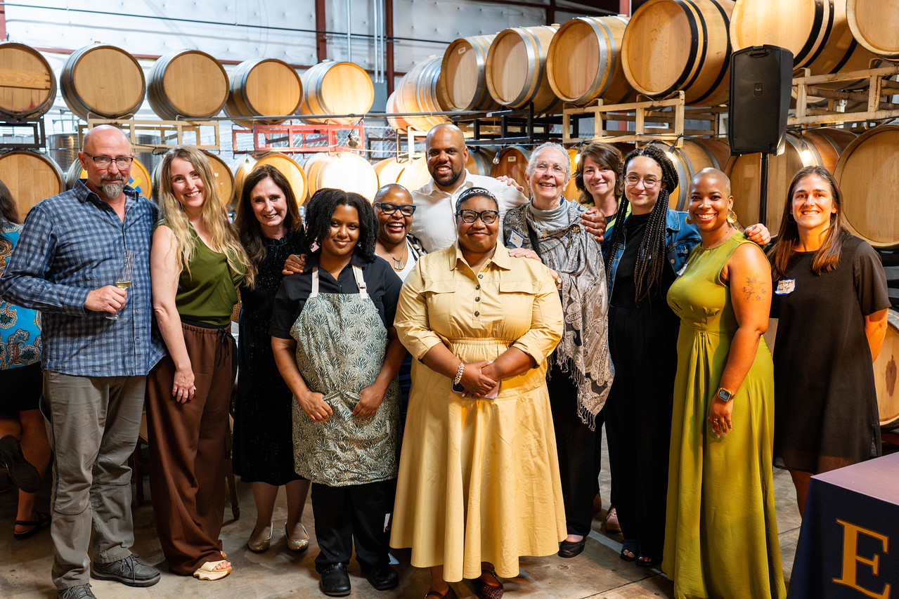 Group of diverse people standing together indoors, smiling, with barrels and industrial background behind them.