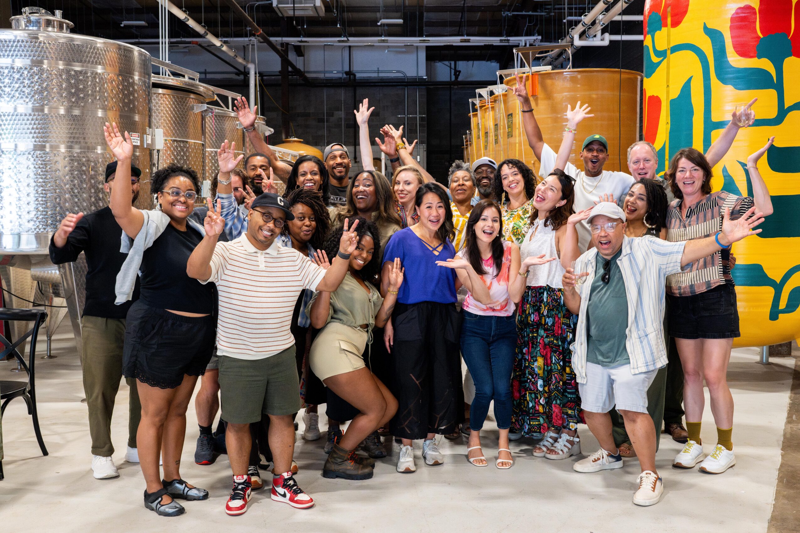 Group of diverse people smiling and waving indoors with colorful artwork and industrial tanks in background.