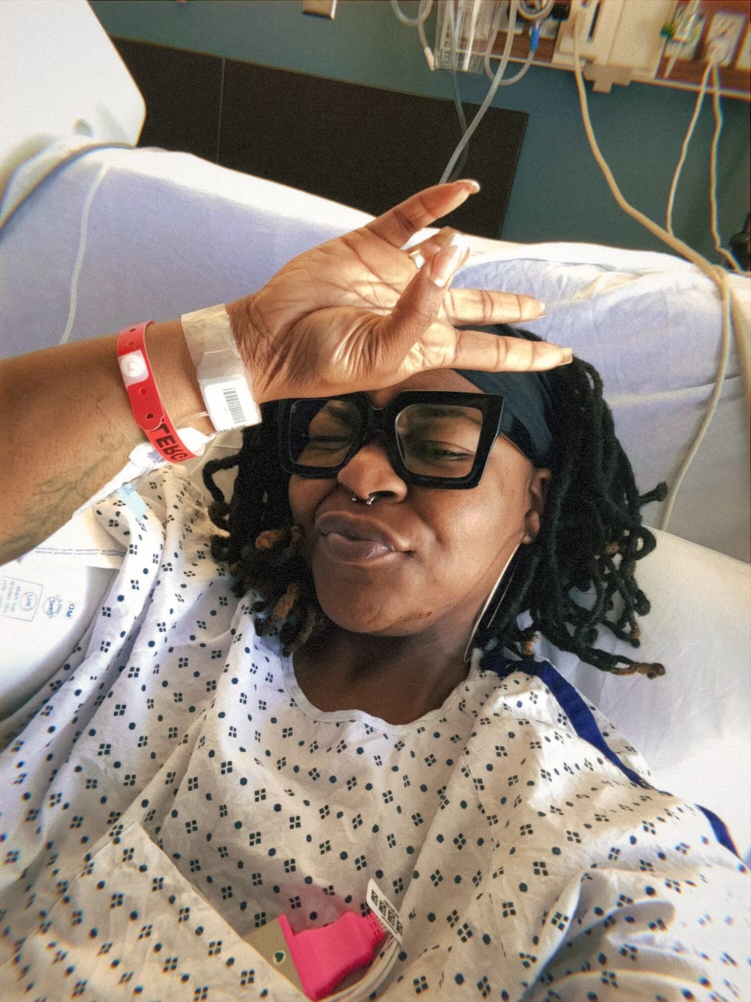Woman in hospital bed making a peace sign with her hand, wearing glasses and hospital gown.