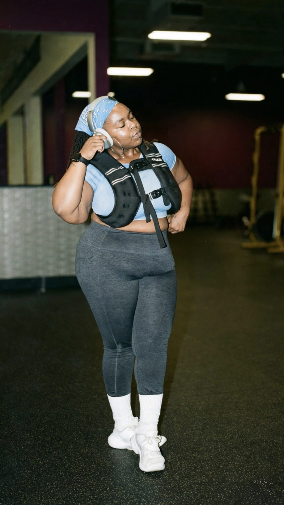 Woman standing in a gym, wearing workout clothes and a life jacket, with her hand on her head, looking to the side.