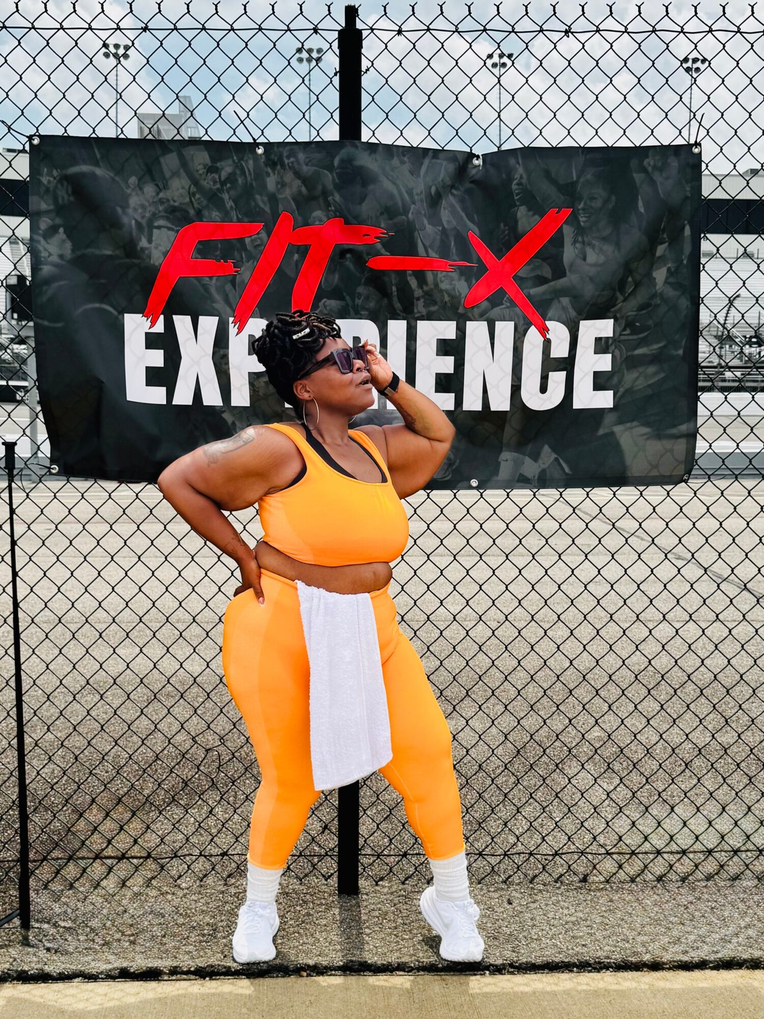 Woman in orange workout outfit standing in front of a chain-link fence with a black banner reading 'FIT-EXPERIENCE'.
