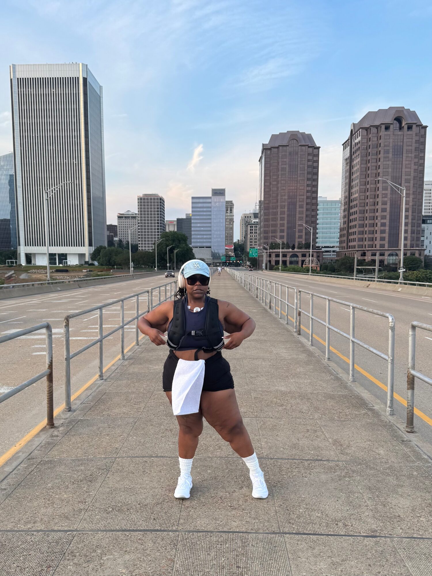Person standing on a bridge with city buildings in the background, wearing a cap, sunglasses, and athletic clothing.