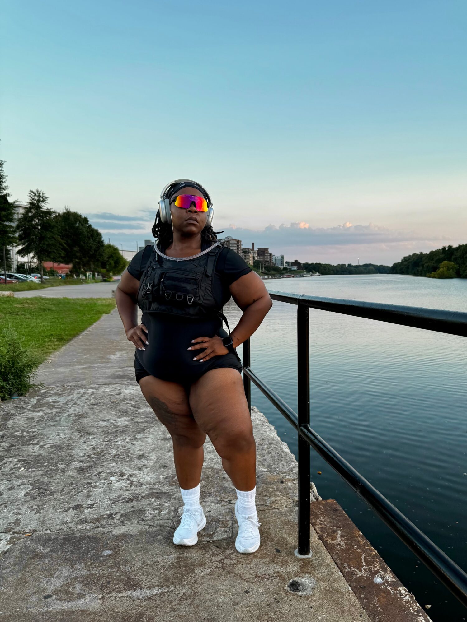 Person standing on a riverside path wearing a black outfit, white shoes, and sunglasses, with a railing beside the water.