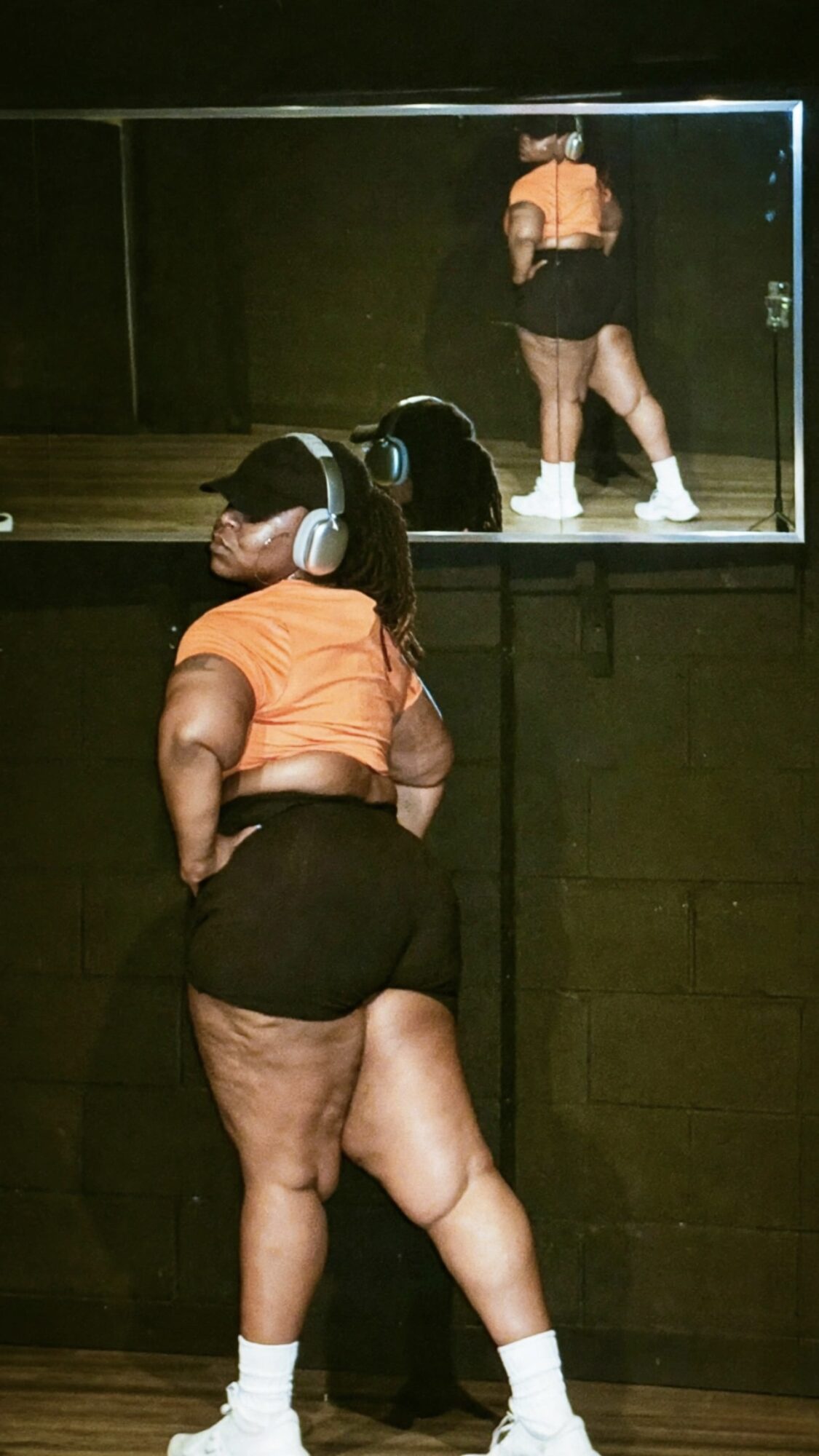 Person with headphones and orange shirt looking at a mirror reflecting a person in black shorts and white shoes.