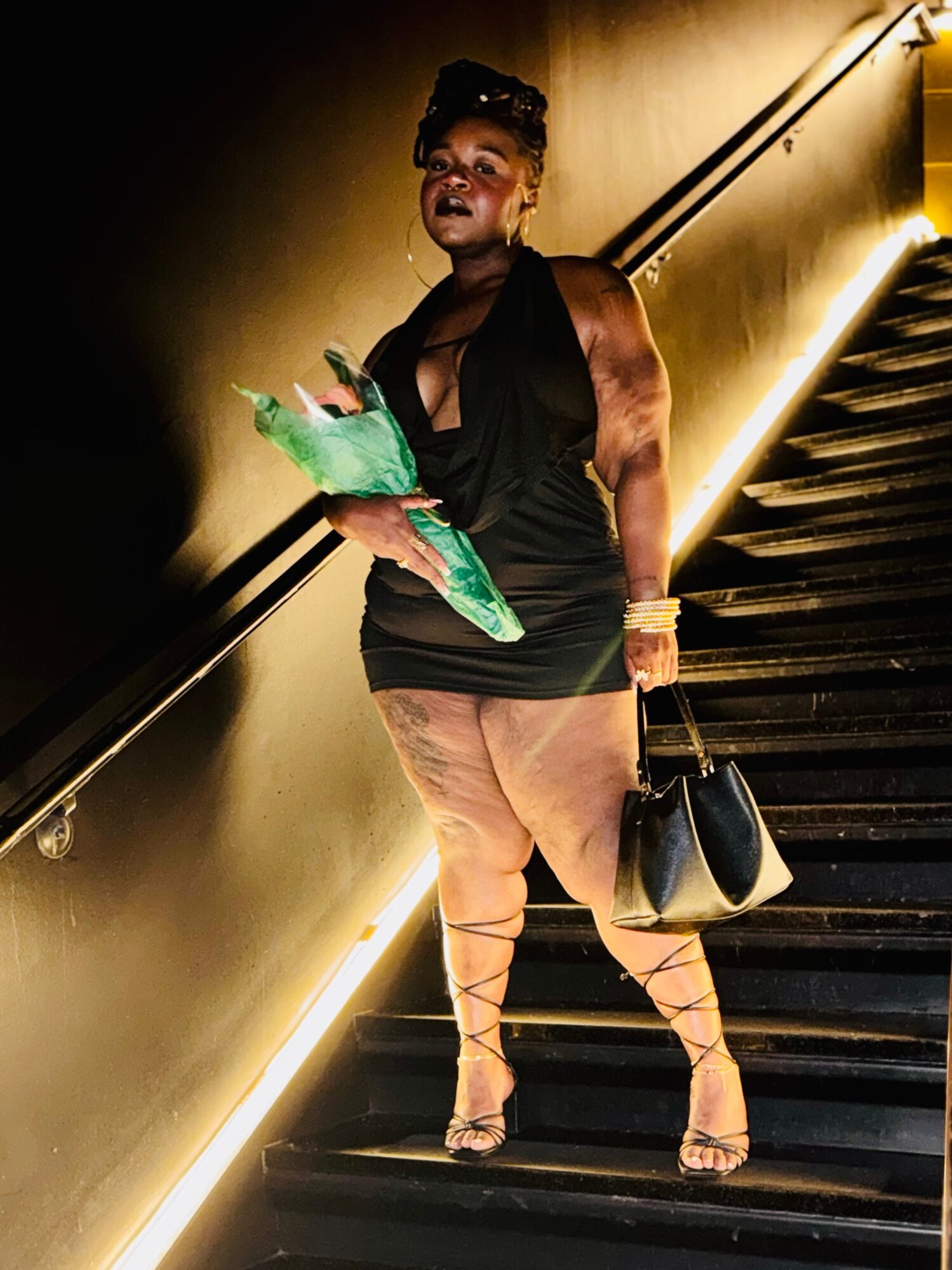 Woman standing on stairs holding a green object and a black handbag, illuminated by warm lighting, dark background.