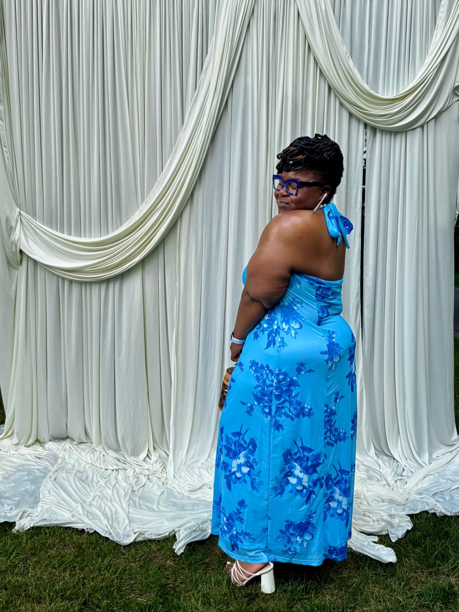 Person in a blue floral dress standing with back to camera, facing white draped curtain backdrop, outdoors on grass