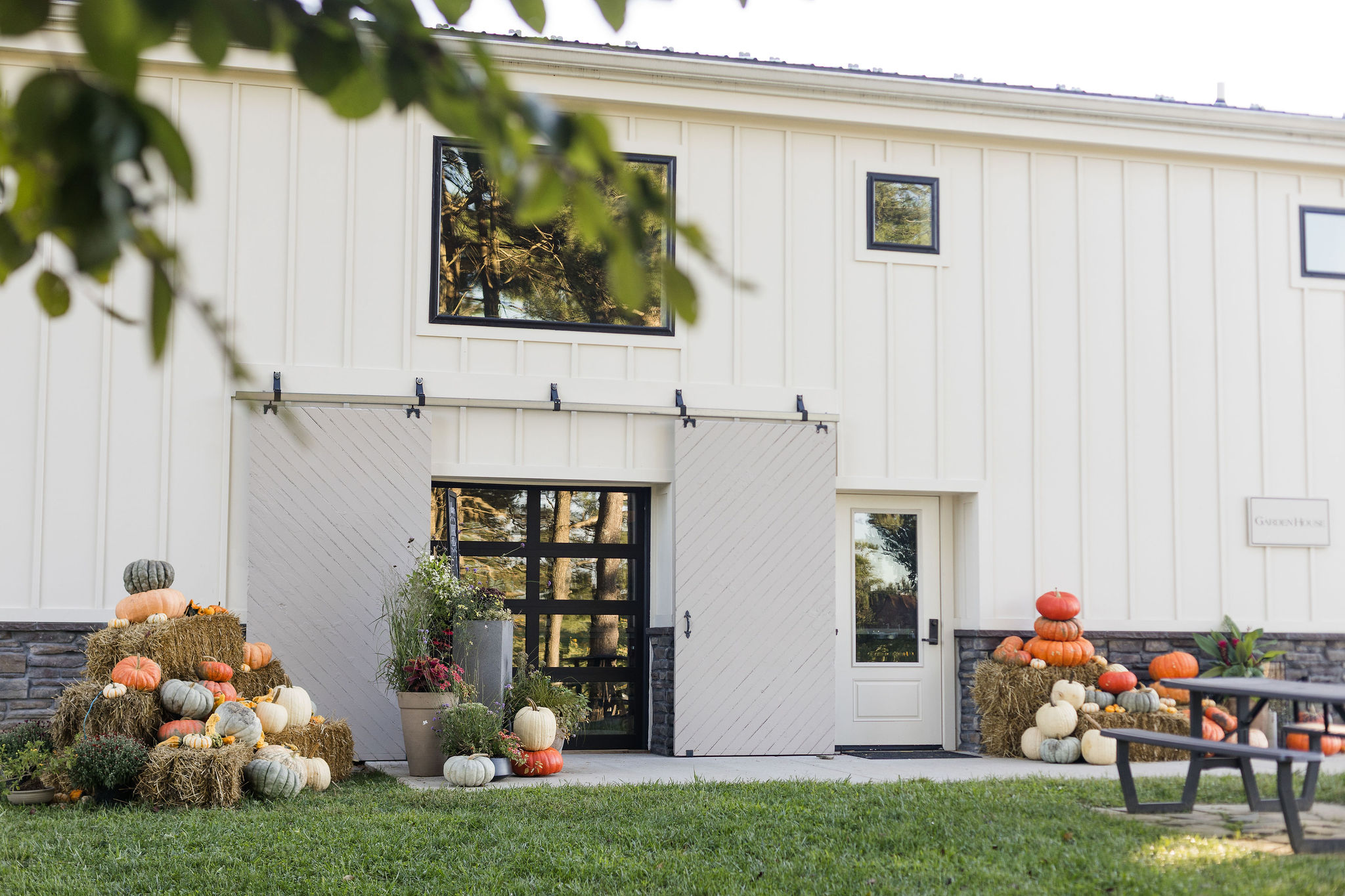 White building with black door, surrounded by stacked pumpkins and hay bales, outdoor seating, and greenery.