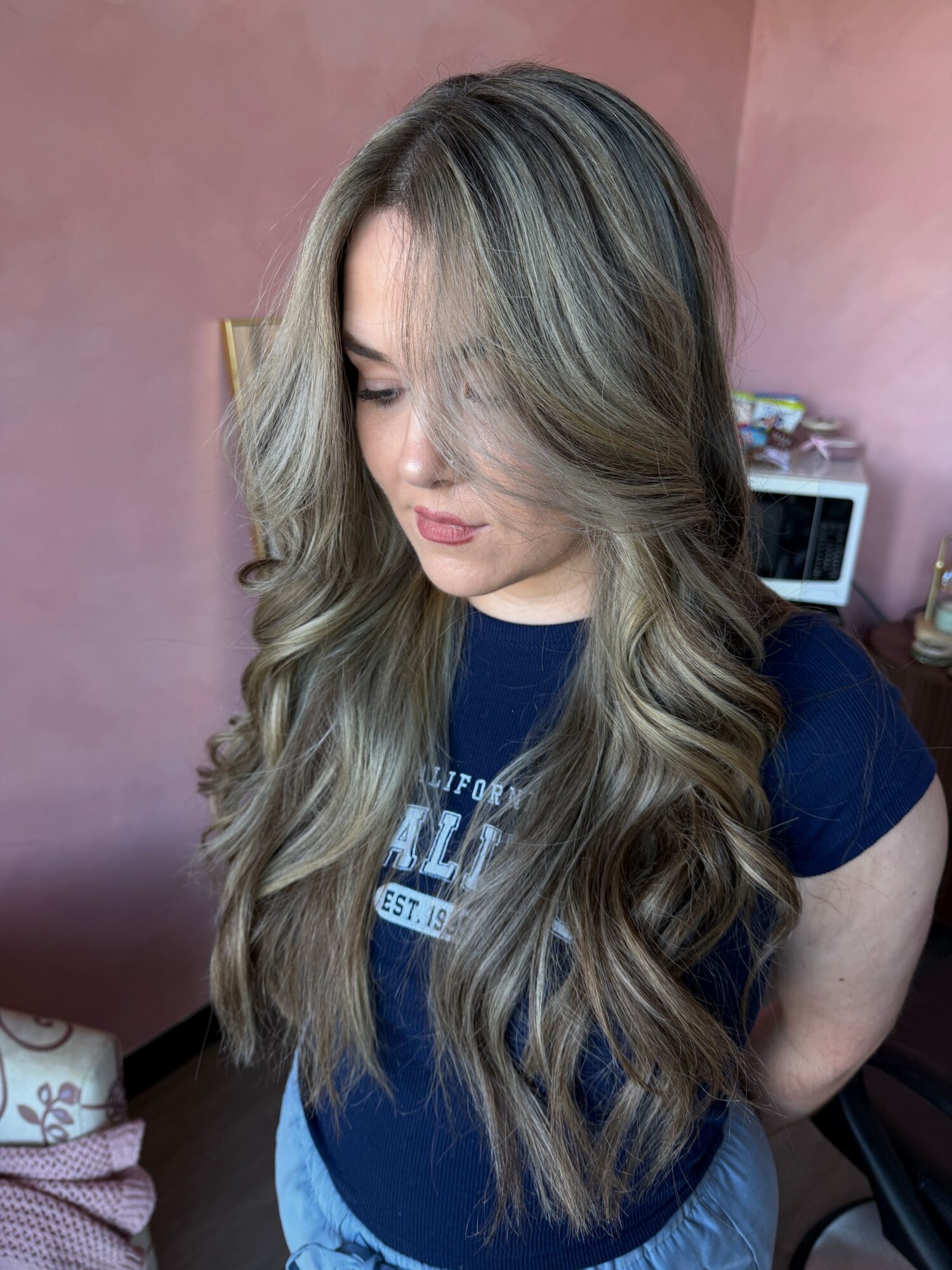 Young woman with long, wavy hair sitting indoors, looking down, wearing a dark shirt and jeans.