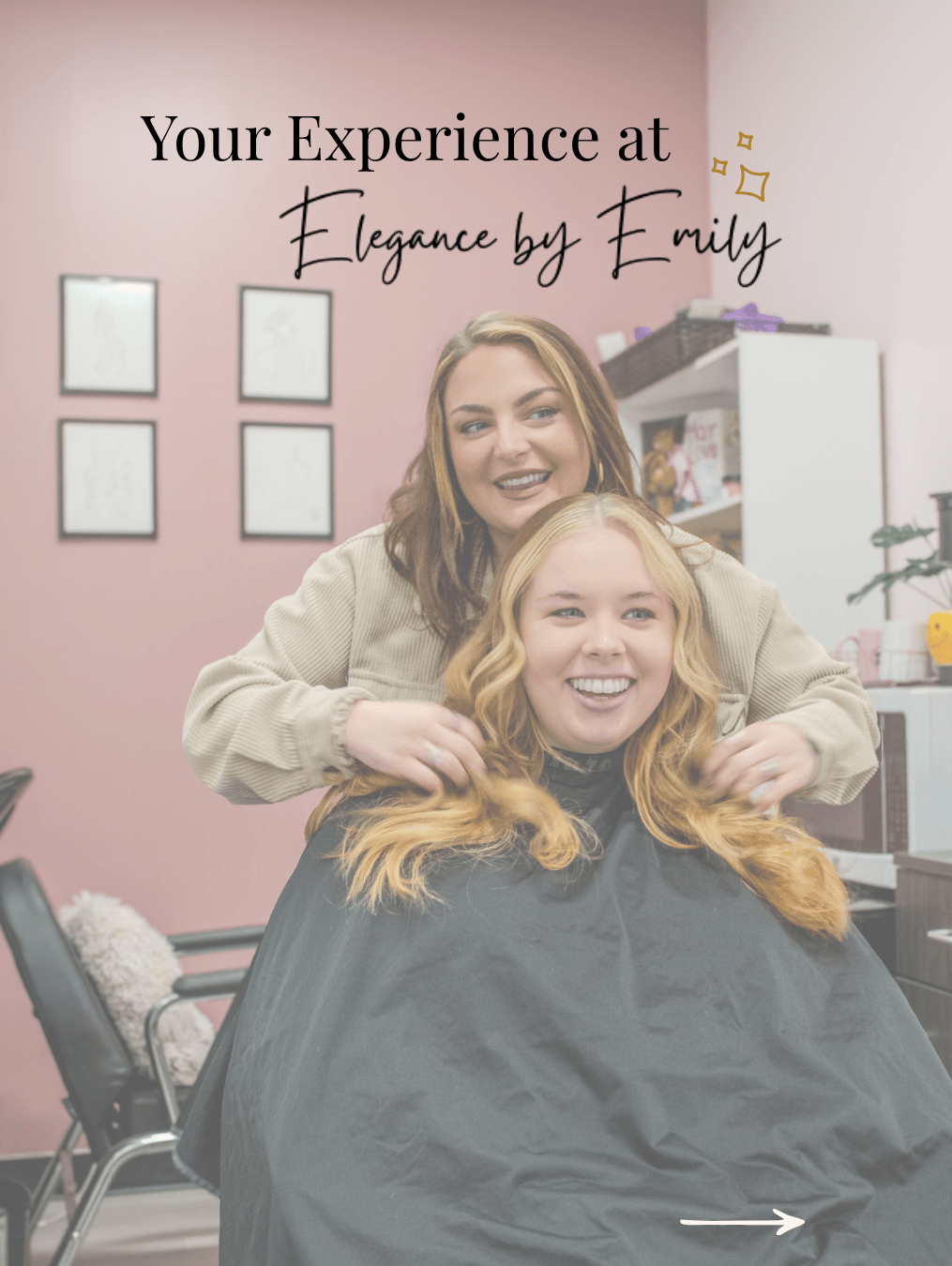 Two women smiling in a salon, one sitting and the other standing behind, with a pink wall and framed pictures in the background.