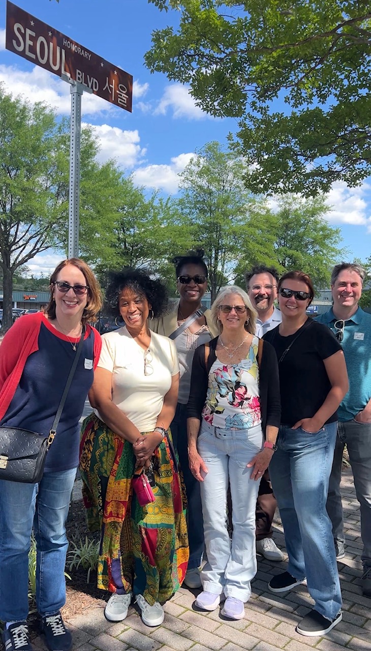 Group of seven diverse people standing outdoors on a sunny day, smiling, with trees and a street sign in the background.