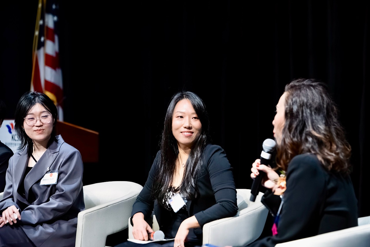 Three women seated on stage, one holding a microphone, engaged in conversation, with a black background and American flag in the background.