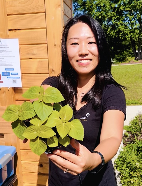 Smiling woman holding a potted plant outdoors, wooden building and green trees in background.