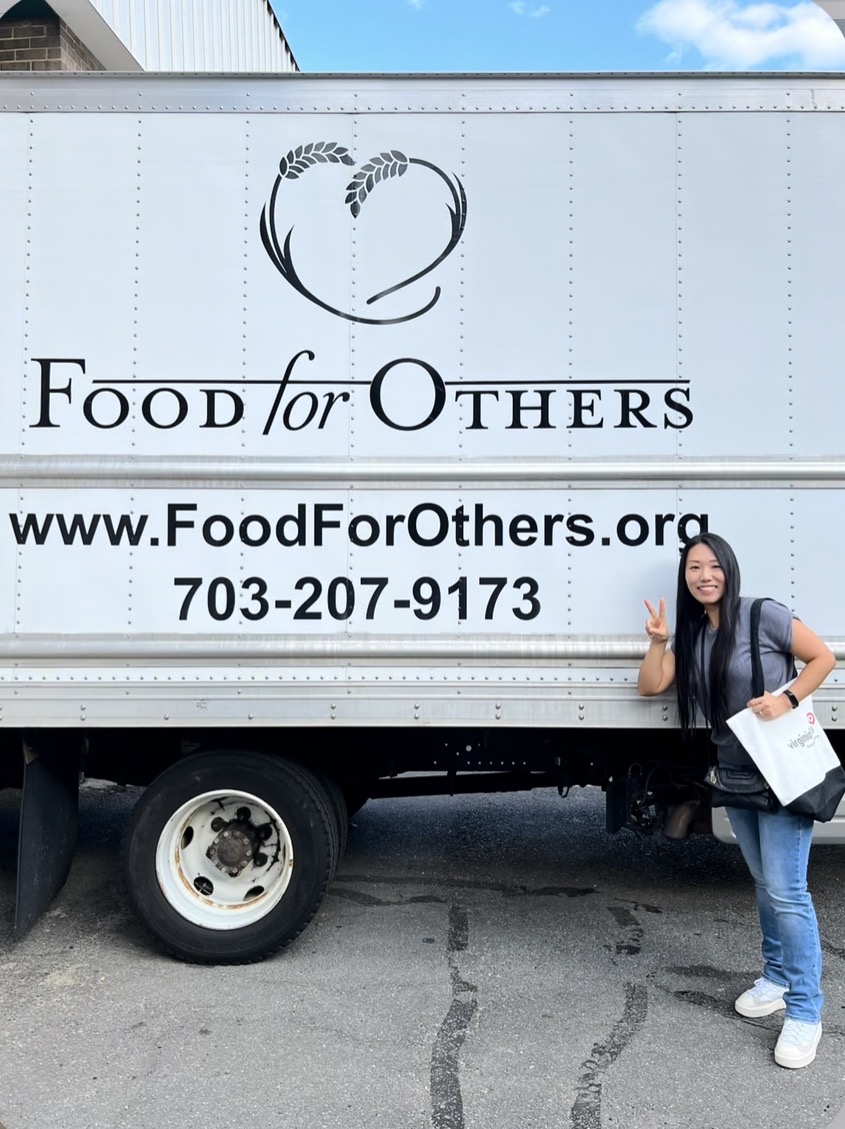 Woman standing next to a food truck with a logo and website, giving a thumbs-up, holding a bag, smiling.
