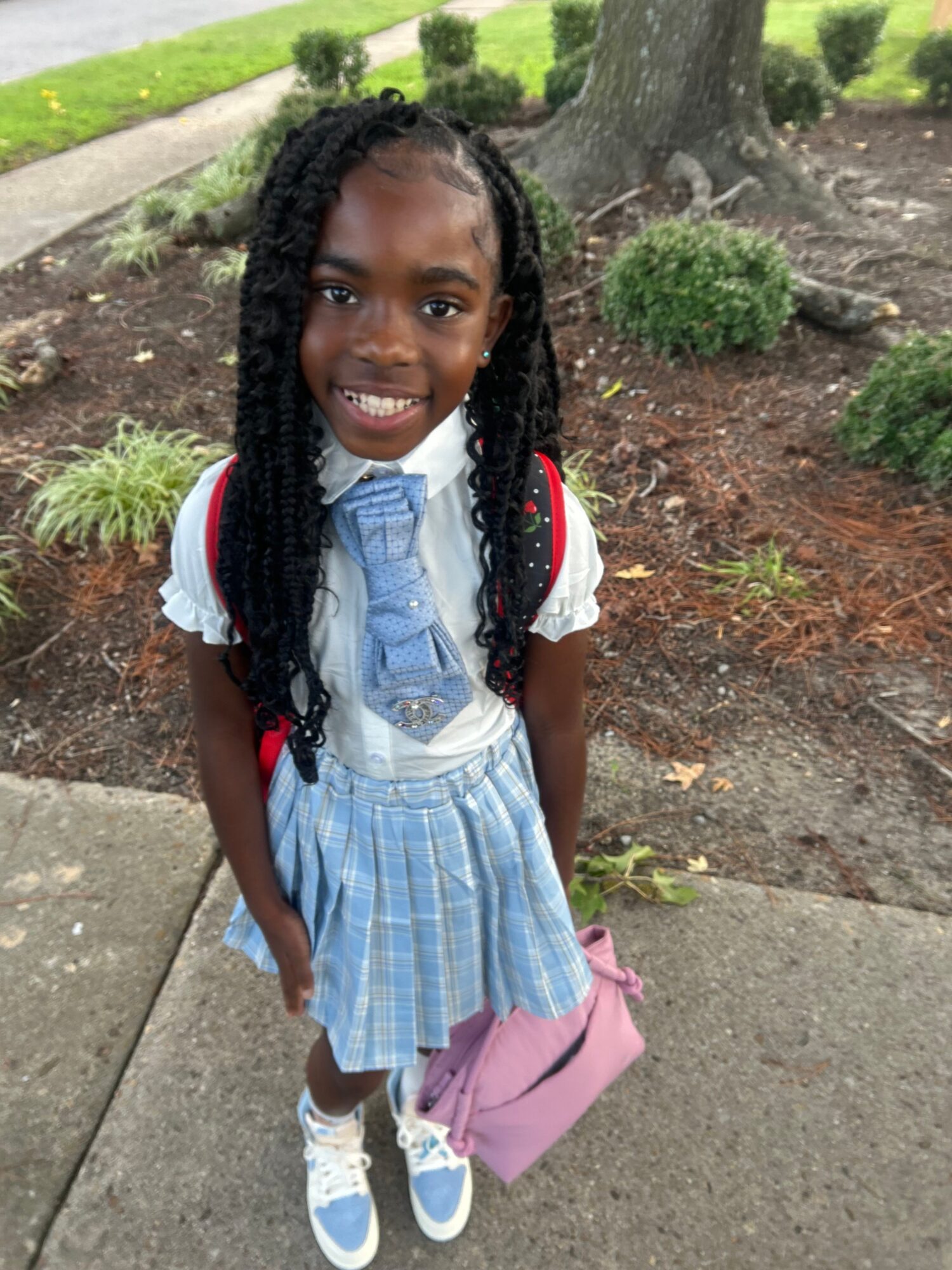 A person with braided hair stands on a sidewalk beside a garden, wearing a blue plaid outfit and carrying a pink backpack.