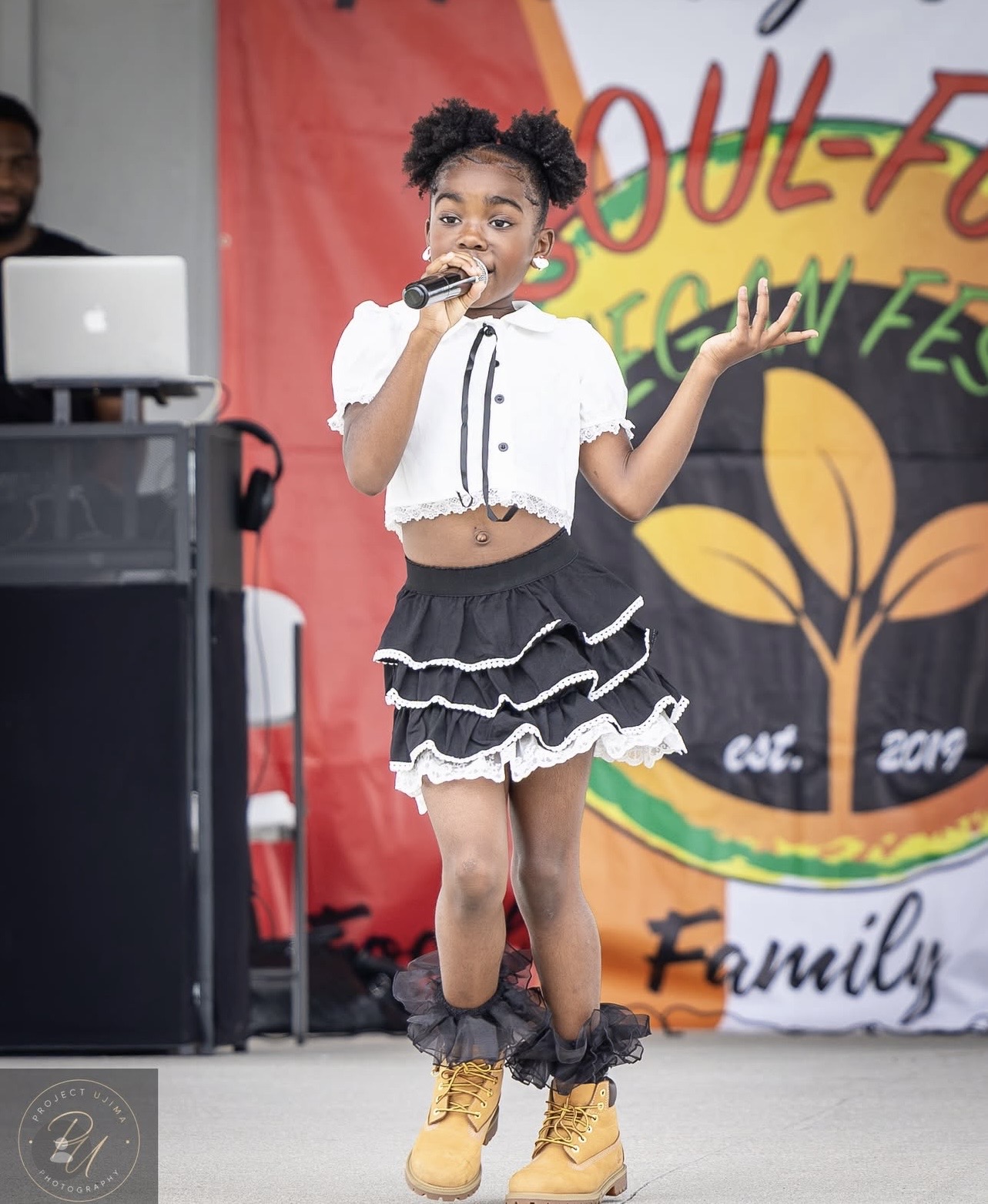 A child sings into a microphone onstage, wearing a white top, black ruffled skirt, and yellow boots.