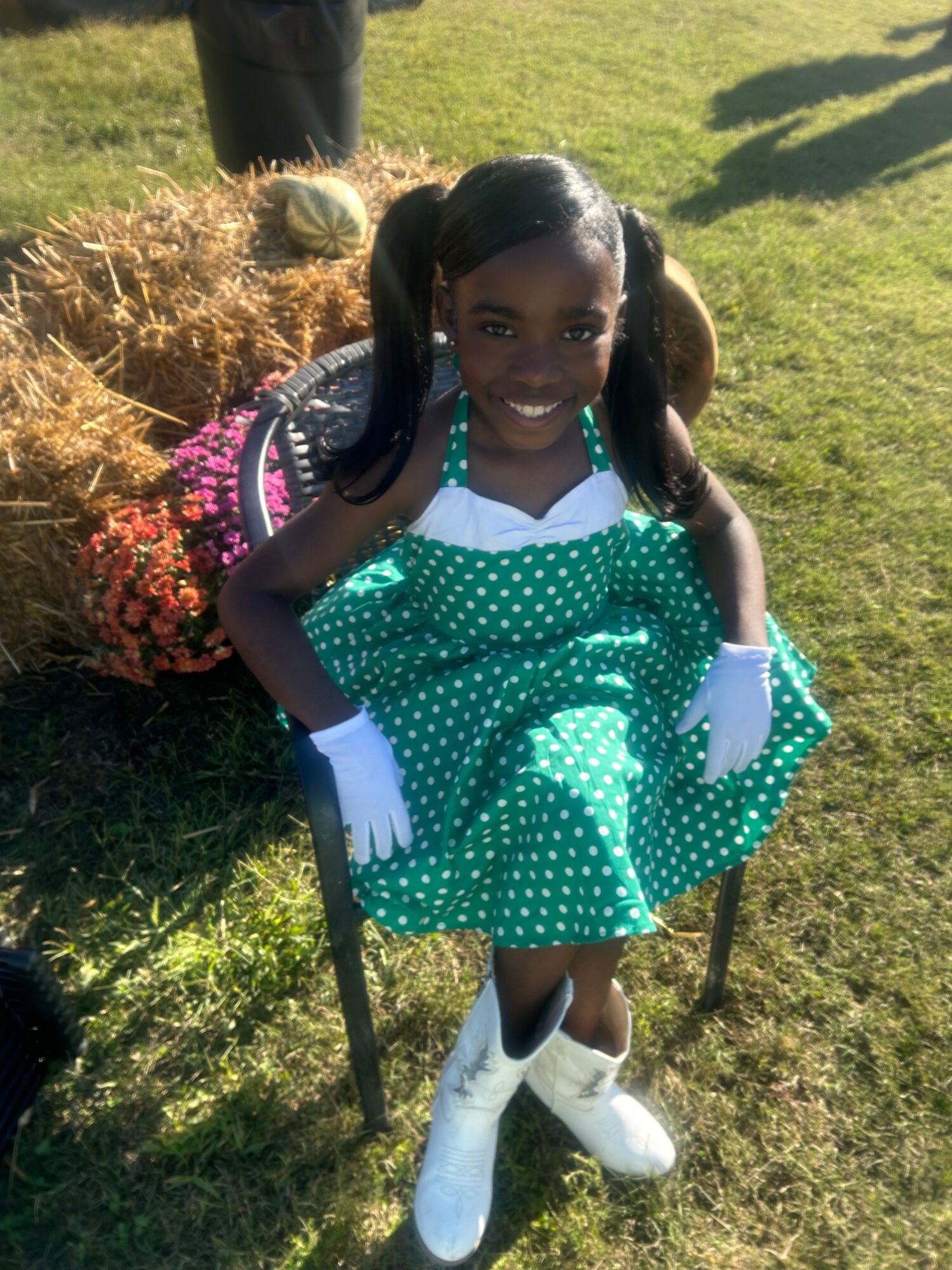 Person in a green polka-dot dress seated in a chair beside flowers on grass.