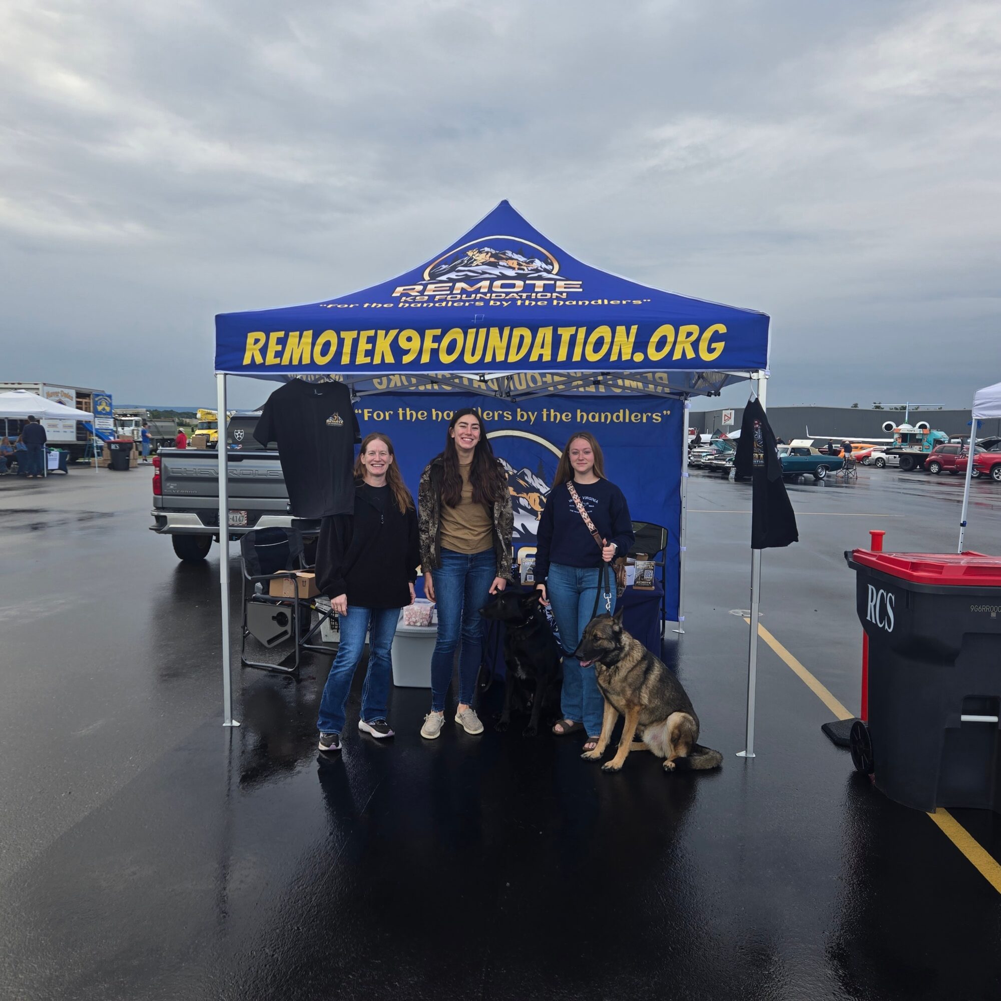 Three women and a dog in front of a blue tent with yellow text on a parking lot, overcast sky.