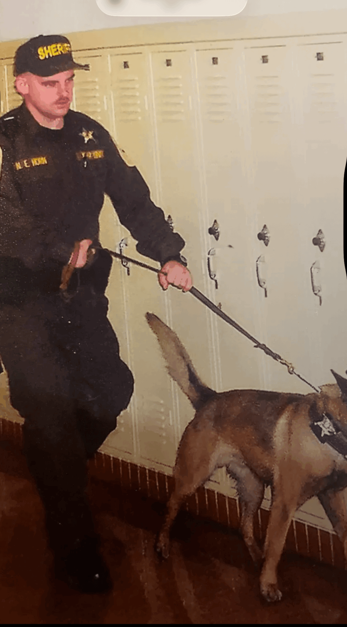 Police officer in uniform with a police dog in a hallway with lockers, holding the dog's leash.