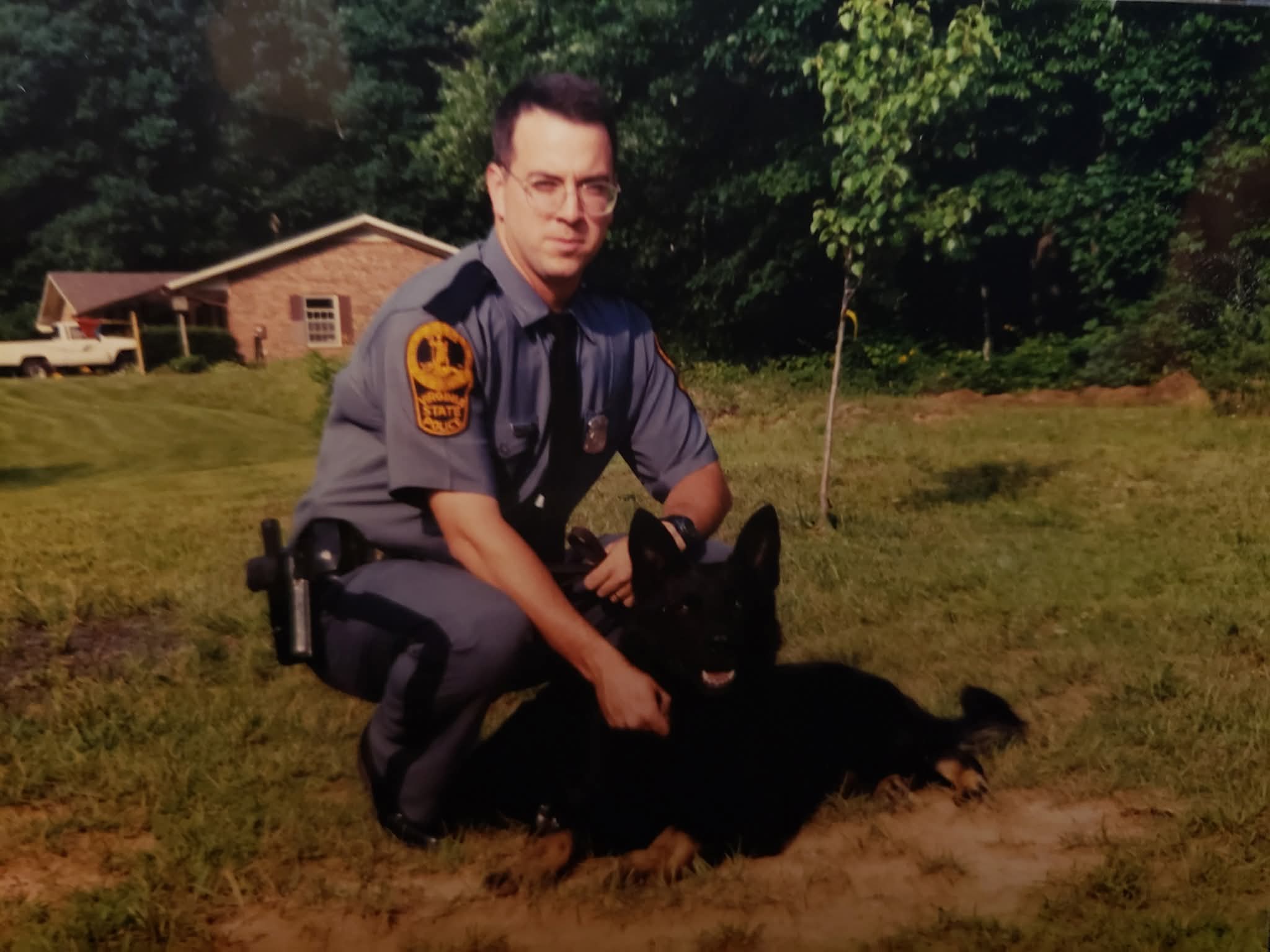 Police officer kneeling on grass with a German Shepherd dog outdoors, trees and a building in background.