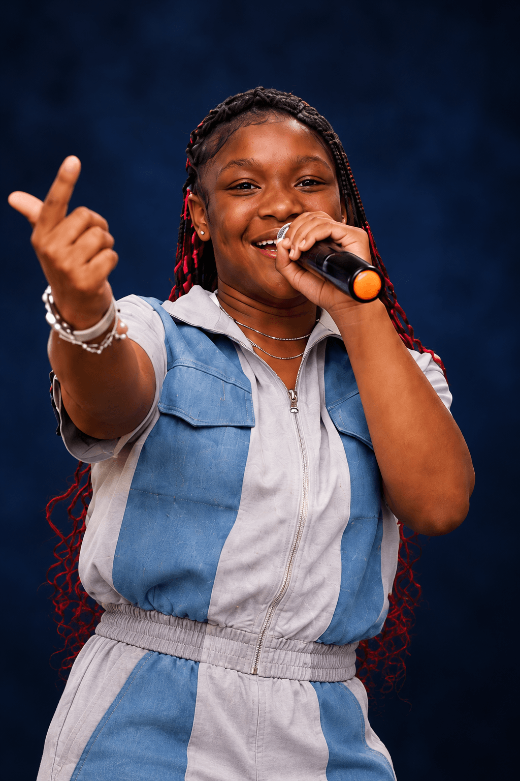 Young girl with braided hair smiling, holding a microphone, and raising her hand in front of a dark background.