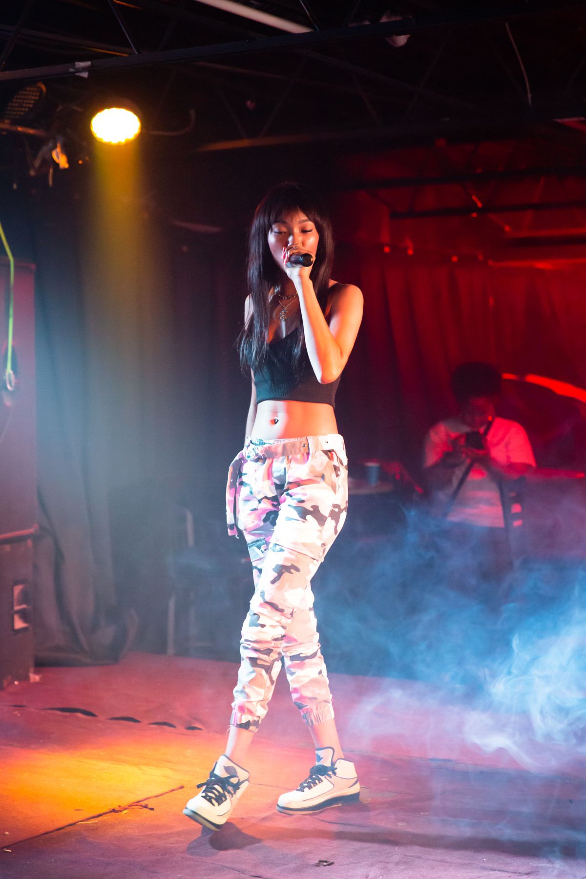 Young woman singing into a microphone on stage with colorful lighting and smoke effects.