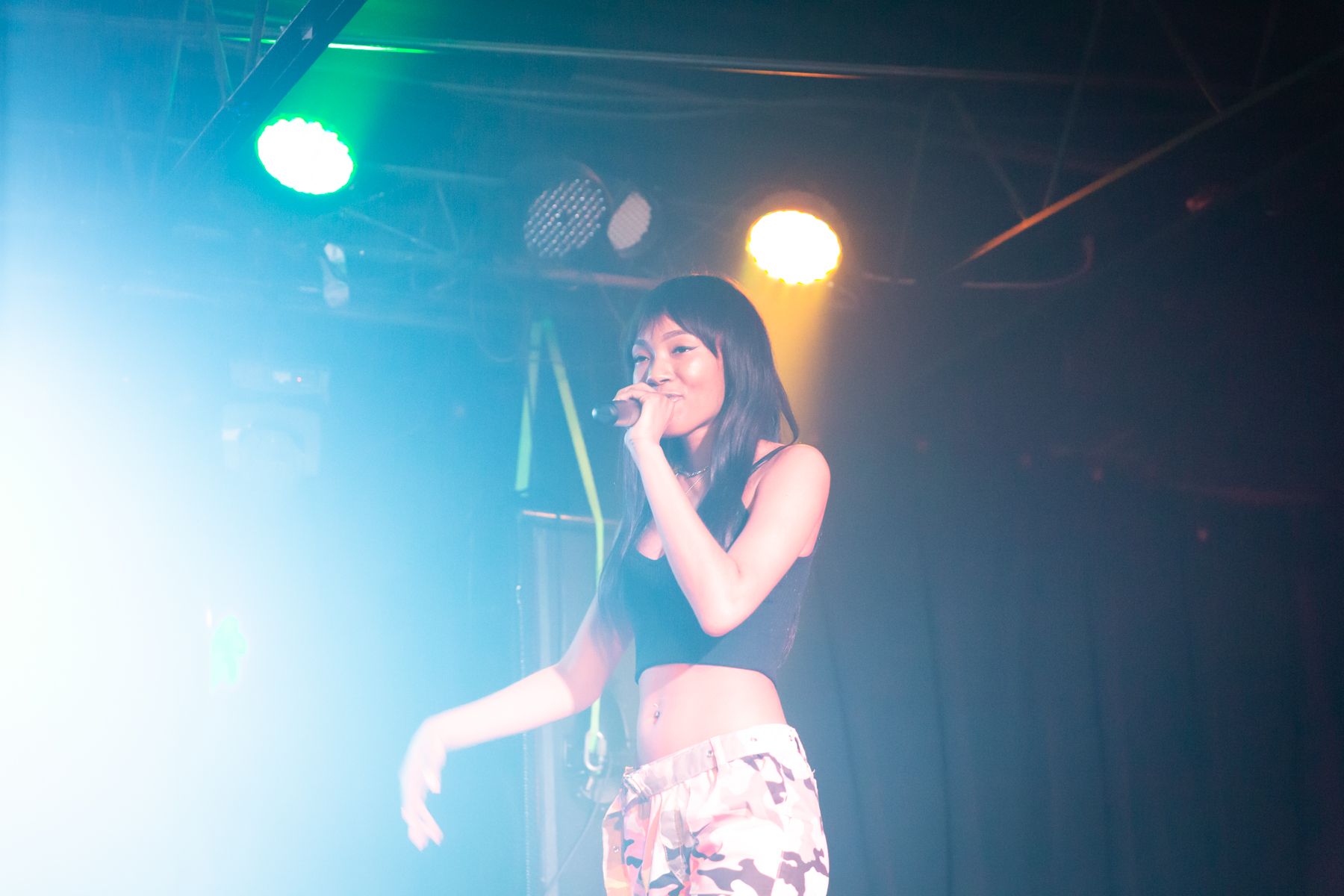 Young woman singing into a microphone on stage with colorful lights, wearing a black crop top and patterned pants.