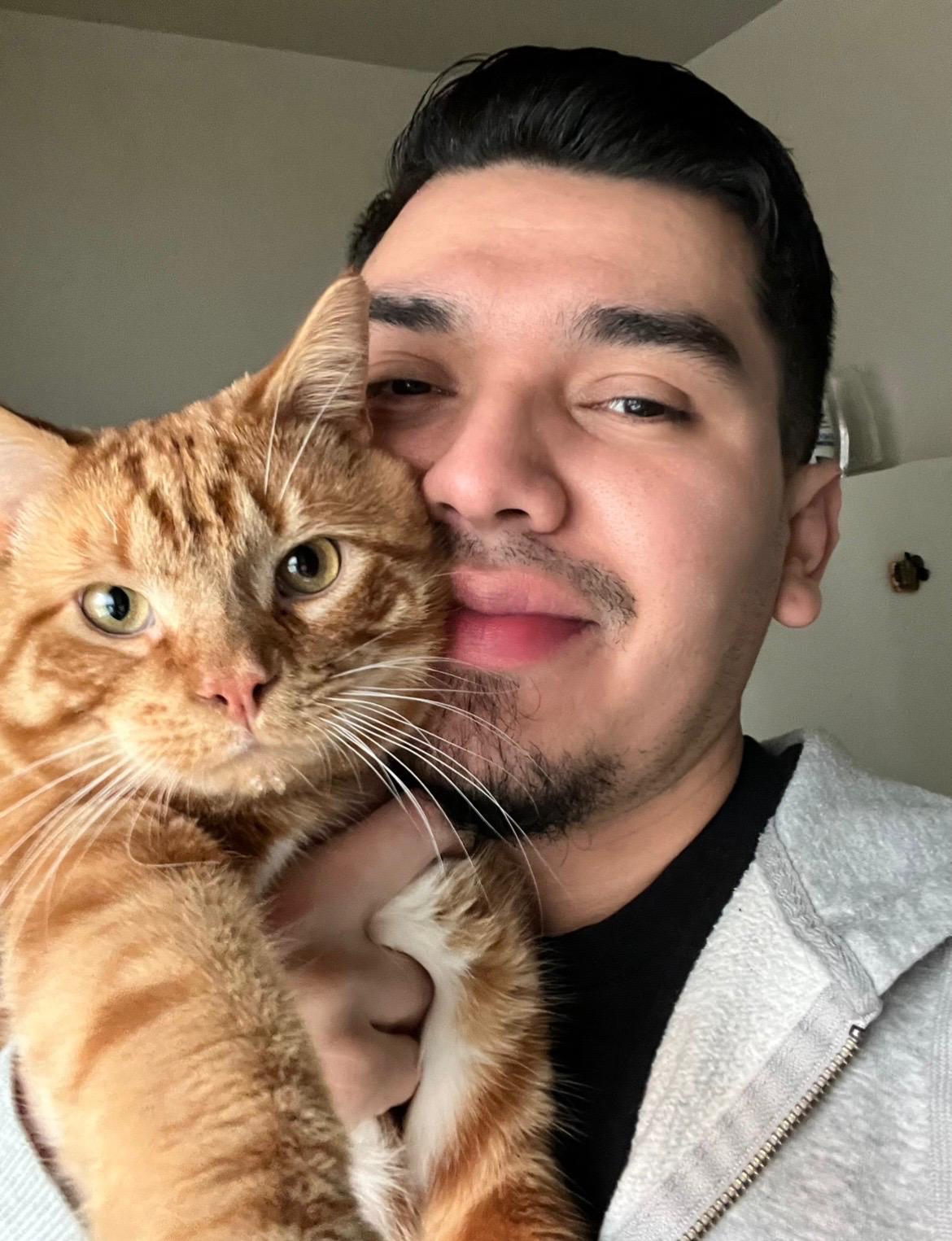Person with dark hair and mustache holding an orange tabby cat close to face, both looking at the camera.