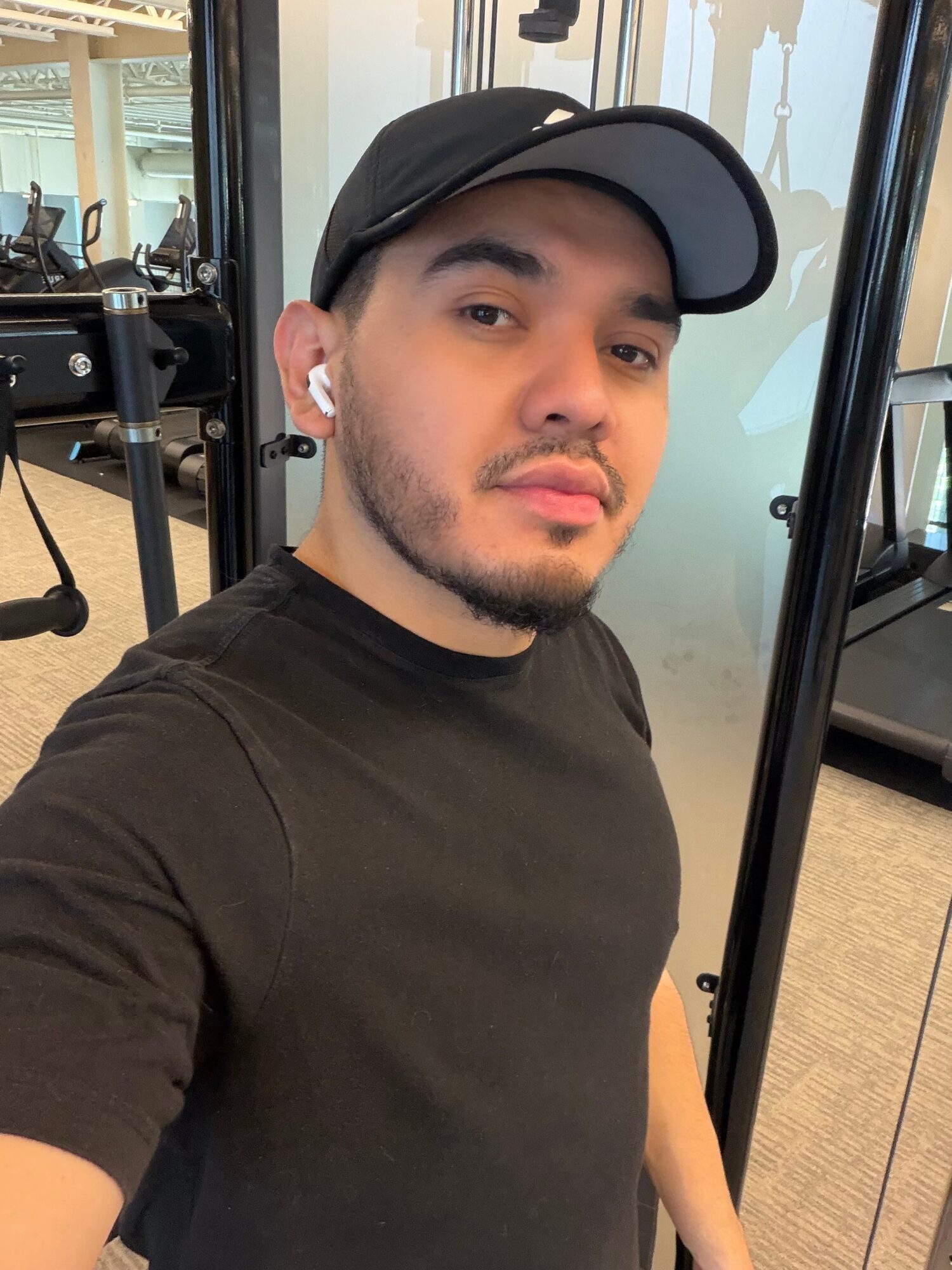 Young man taking a selfie in a gym, wearing a black cap, black t-shirt, and wireless earbuds.
