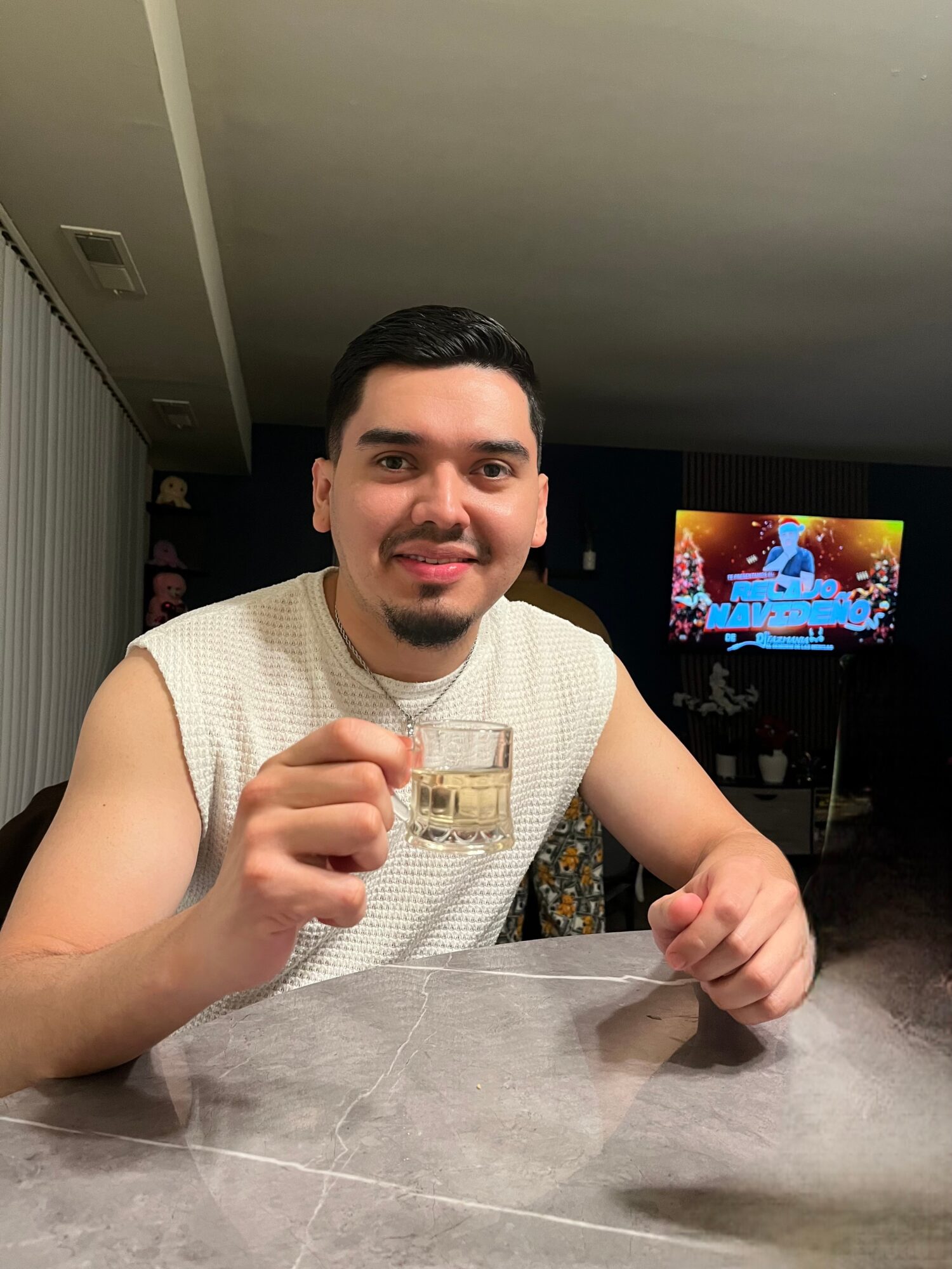 Young man with dark hair and a goatee, smiling, holding a glass, sitting at a table in a room with a TV in the background.