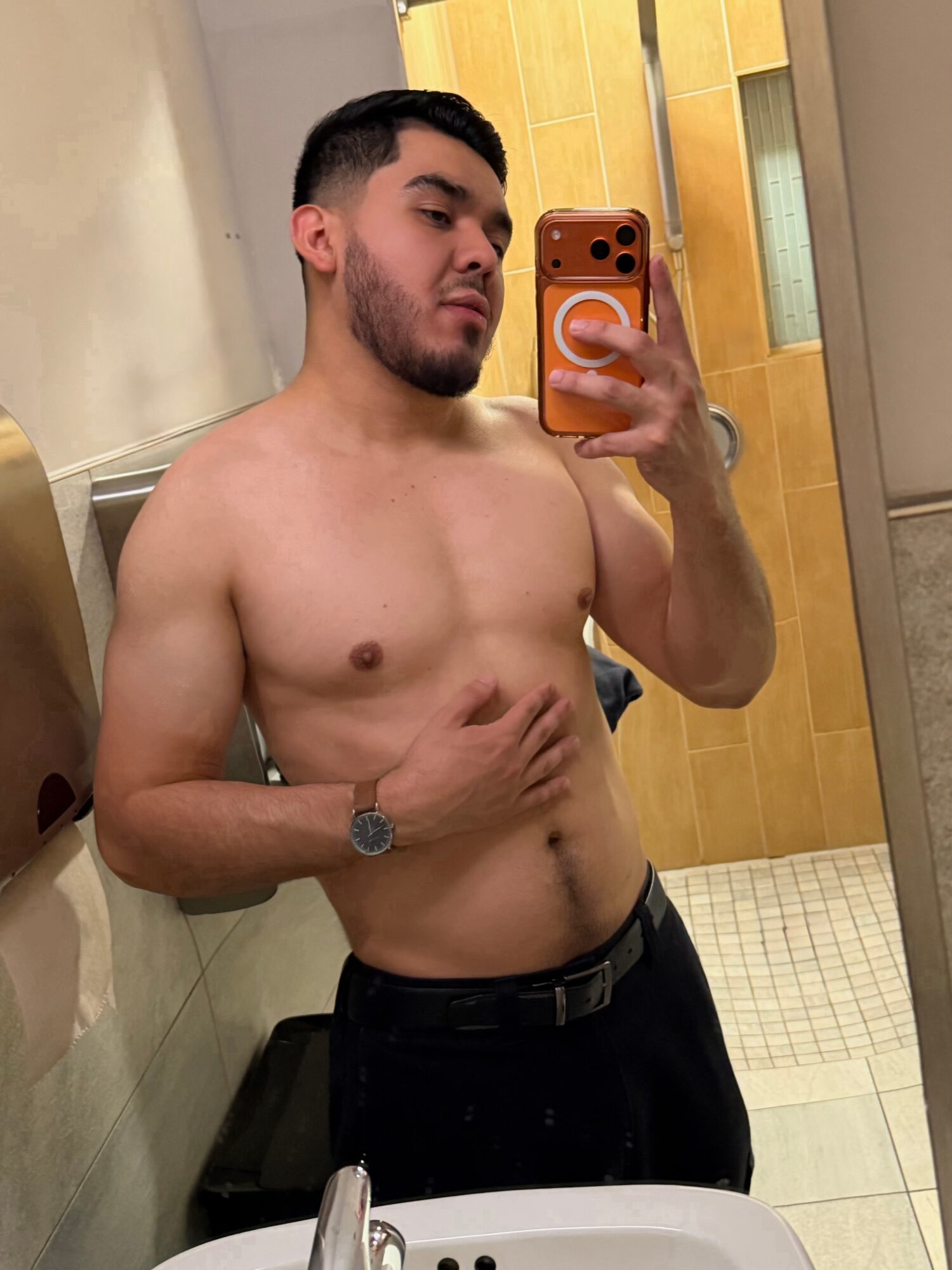 Young man taking a mirror selfie in a bathroom, shirtless, with a neutral expression, holding phone with orange case, standing near a sink.