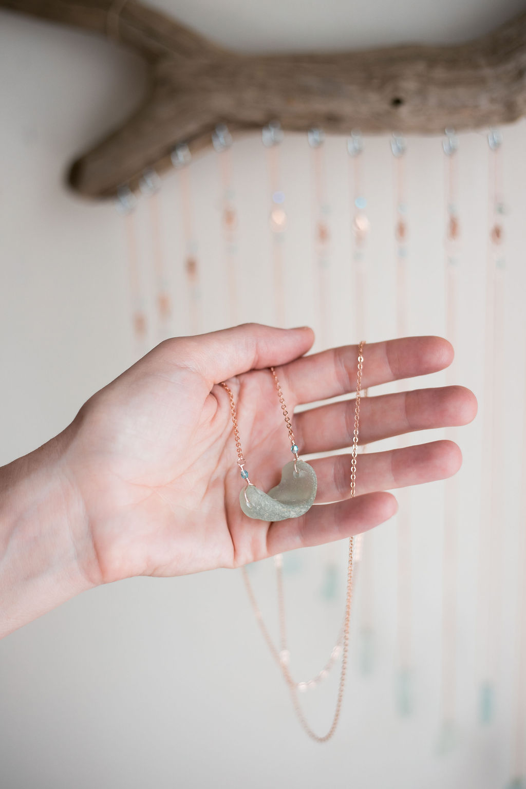 Hand holding a necklace with a moon-shaped pendant, background with hanging beads and a tree branch, neutral lighting.