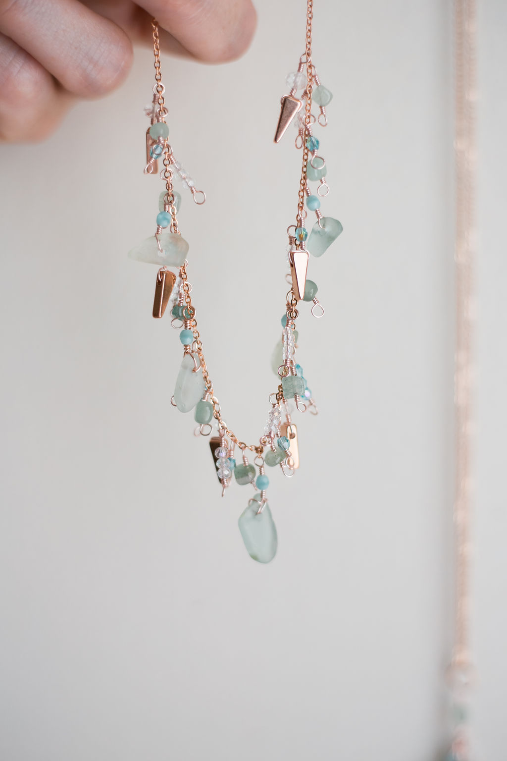 Necklace with sea glass and small beads hanging from a thin chain, held by a person's hand, against a plain background.