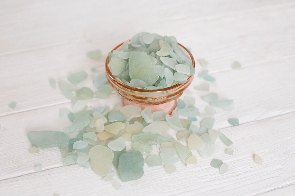 Small copper bowl filled with pastel-colored sea glass pieces on a white wooden surface, with some glass scattered around.