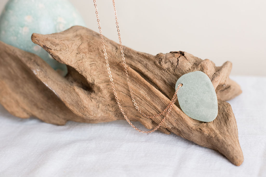 A light blue heart-shaped pendant on a gold chain rests on a piece of driftwood, with a blue sphere in the background.