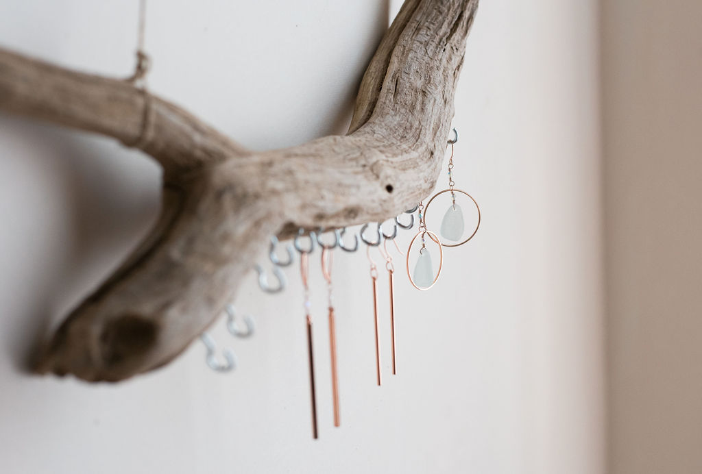 Earrings hanging from a piece of driftwood mounted on a wall, with a neutral background.