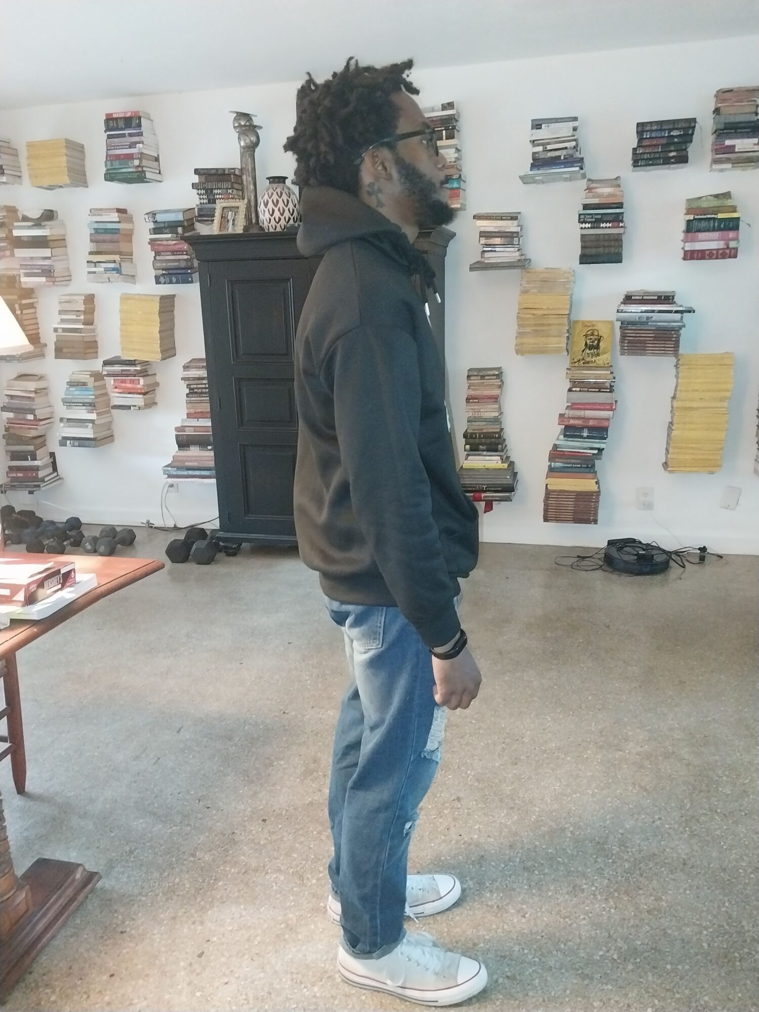 A person stands in profile in front of bookshelves, wearing a dark jacket, blue jeans, and sneakers.