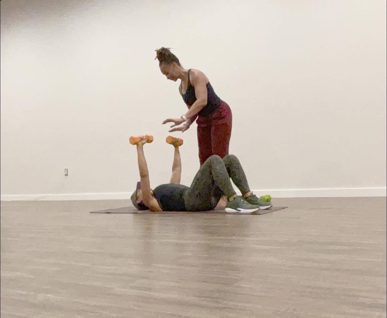 Two women in a dance studio, one lying on the floor with arms raised, the other standing over her, gesturing.