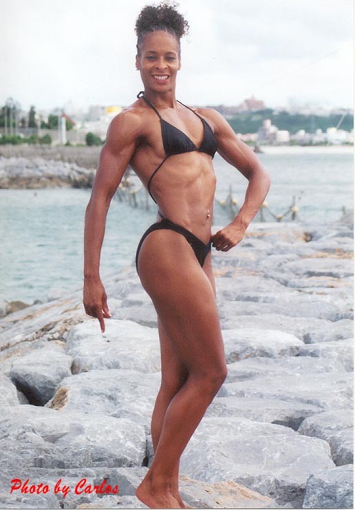 Woman in a black bikini standing on rocks by water, smiling, with cityscape in background.