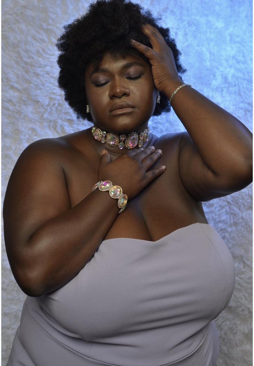 Woman with natural hair, eyes closed, touching her forehead and chest, wearing jewelry, against a textured background.