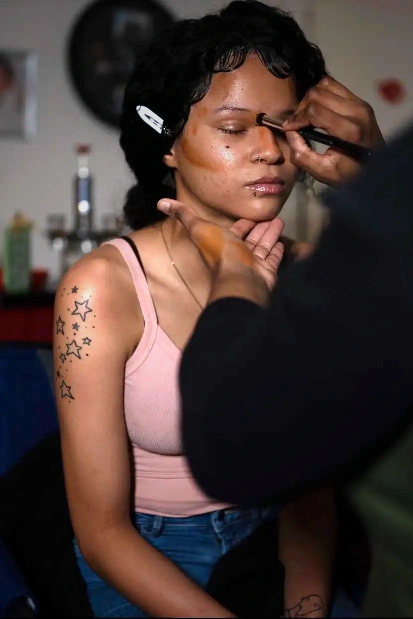 Woman with short dark hair getting makeup applied, wearing a pink tank top, tattoo on her arm, in a room with makeup tools.