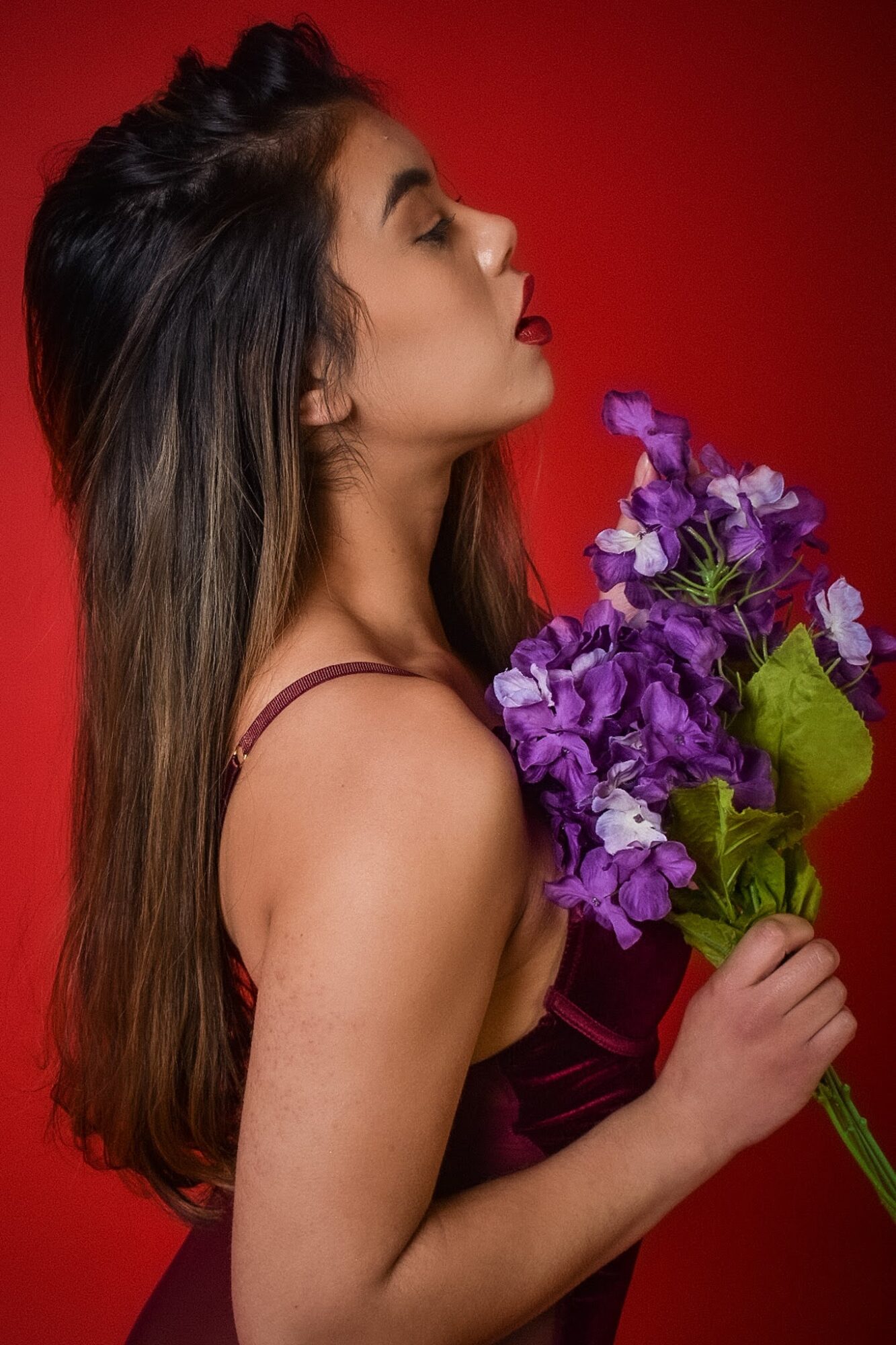 Side profile of a woman with long hair holding a bouquet of purple flowers against a red background.