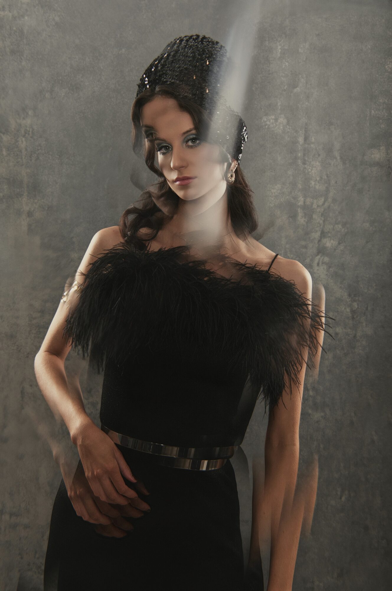 A person in a black dress and beaded headpiece poses against a gray textured backdrop.