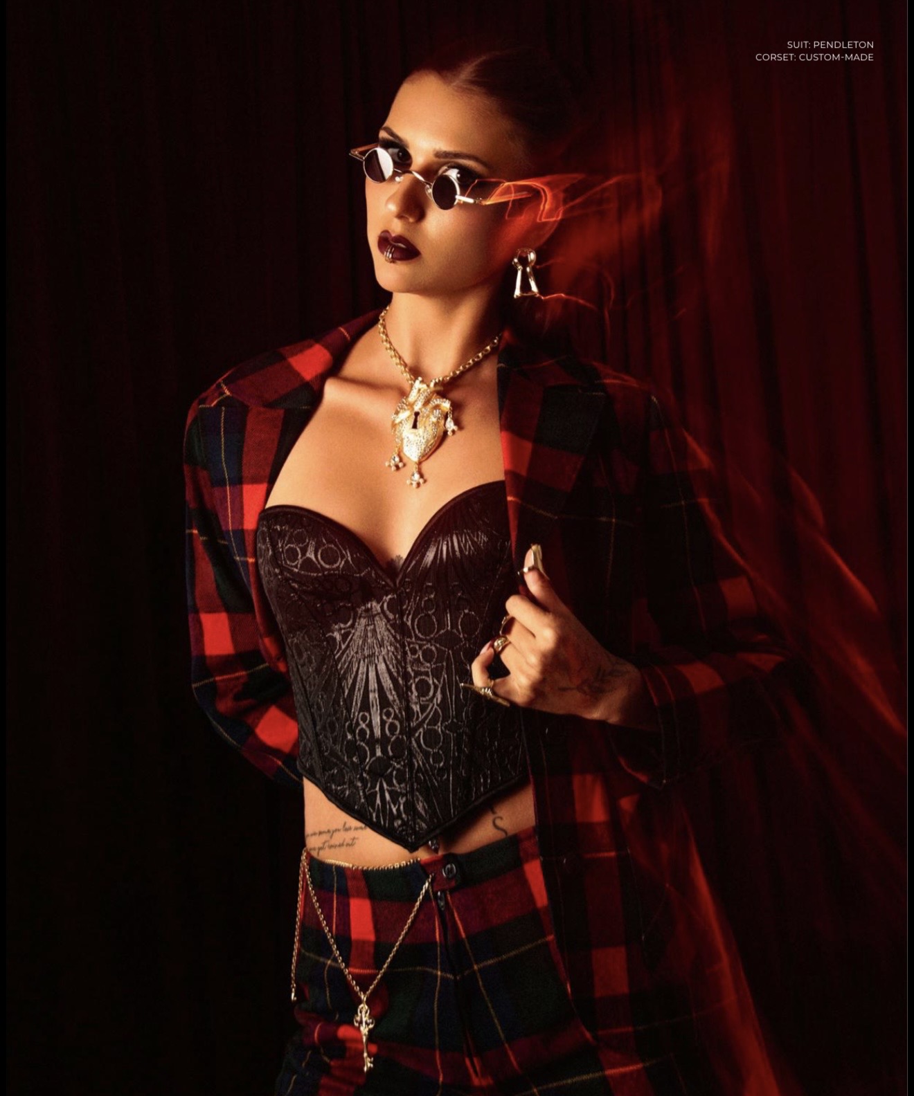 Person wearing sunglasses, a red-and-black plaid outfit, a black corset, and layered necklaces against a dark red backdrop.