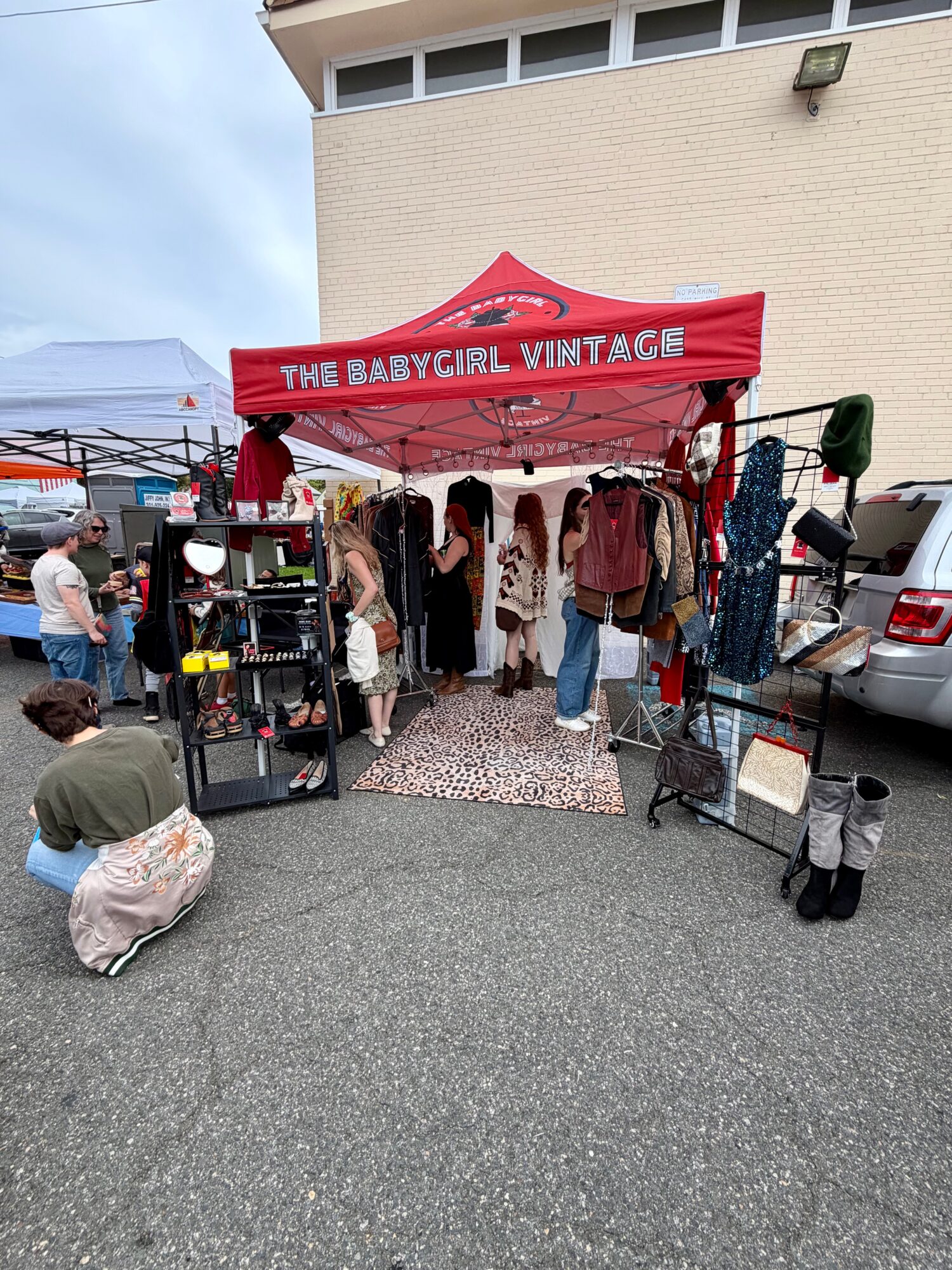Outdoor vintage clothing stall beneath a red canopy, with garments on racks and shoppers nearby.