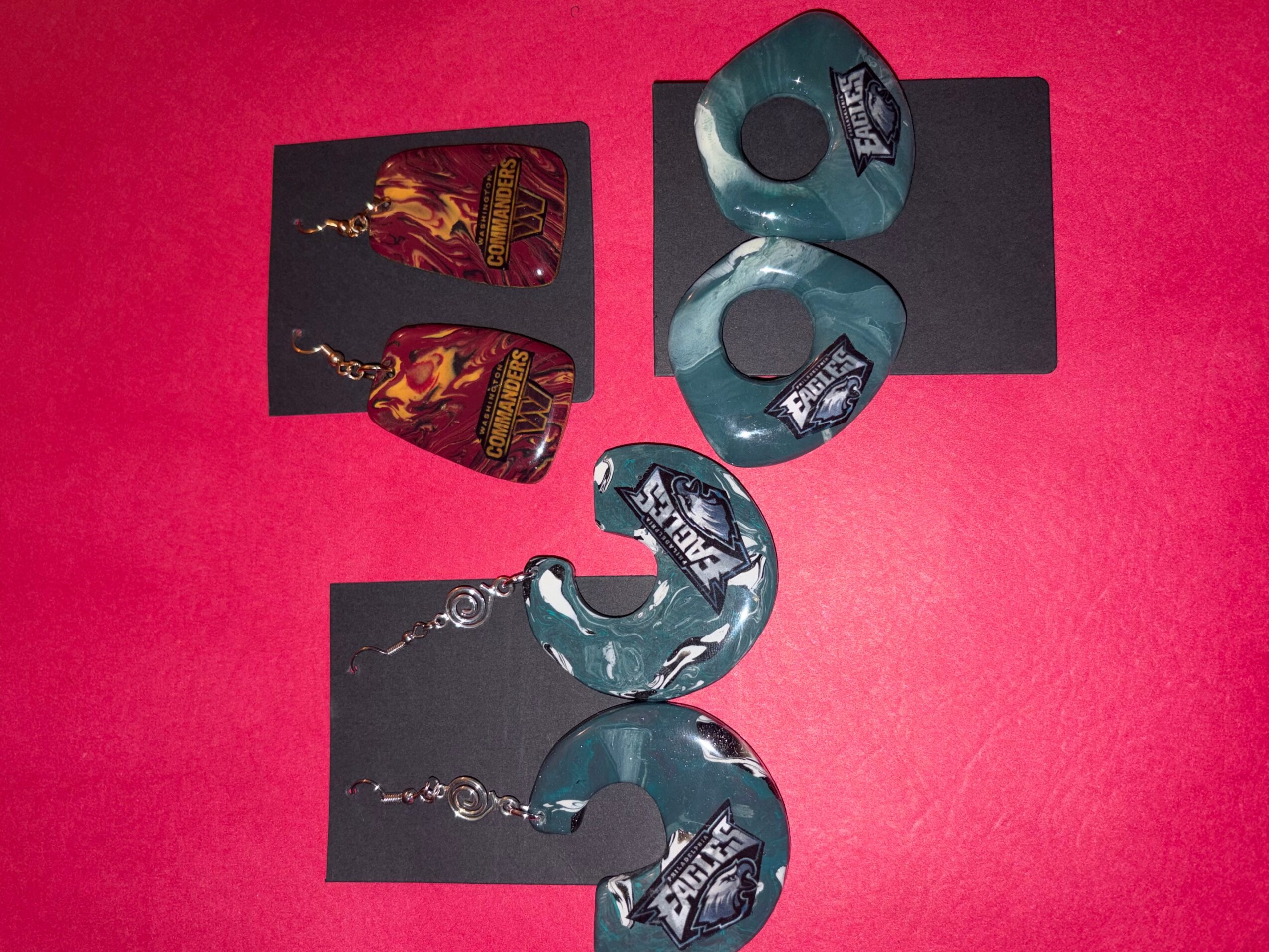 Two earrings and two puzzle pieces on a pink background, with black cards underneath, one earring is blue and the other is red and gold.