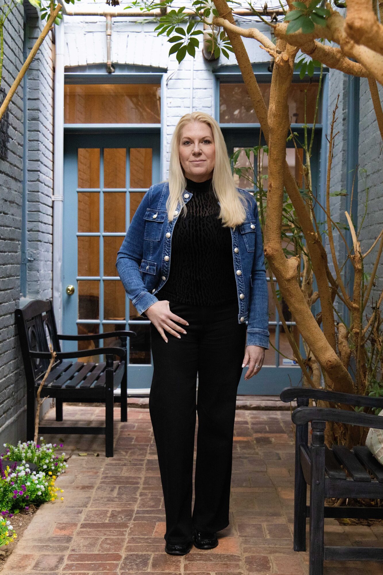 A person stands in a brick courtyard between black benches, with a glass-paneled door and trees behind them.