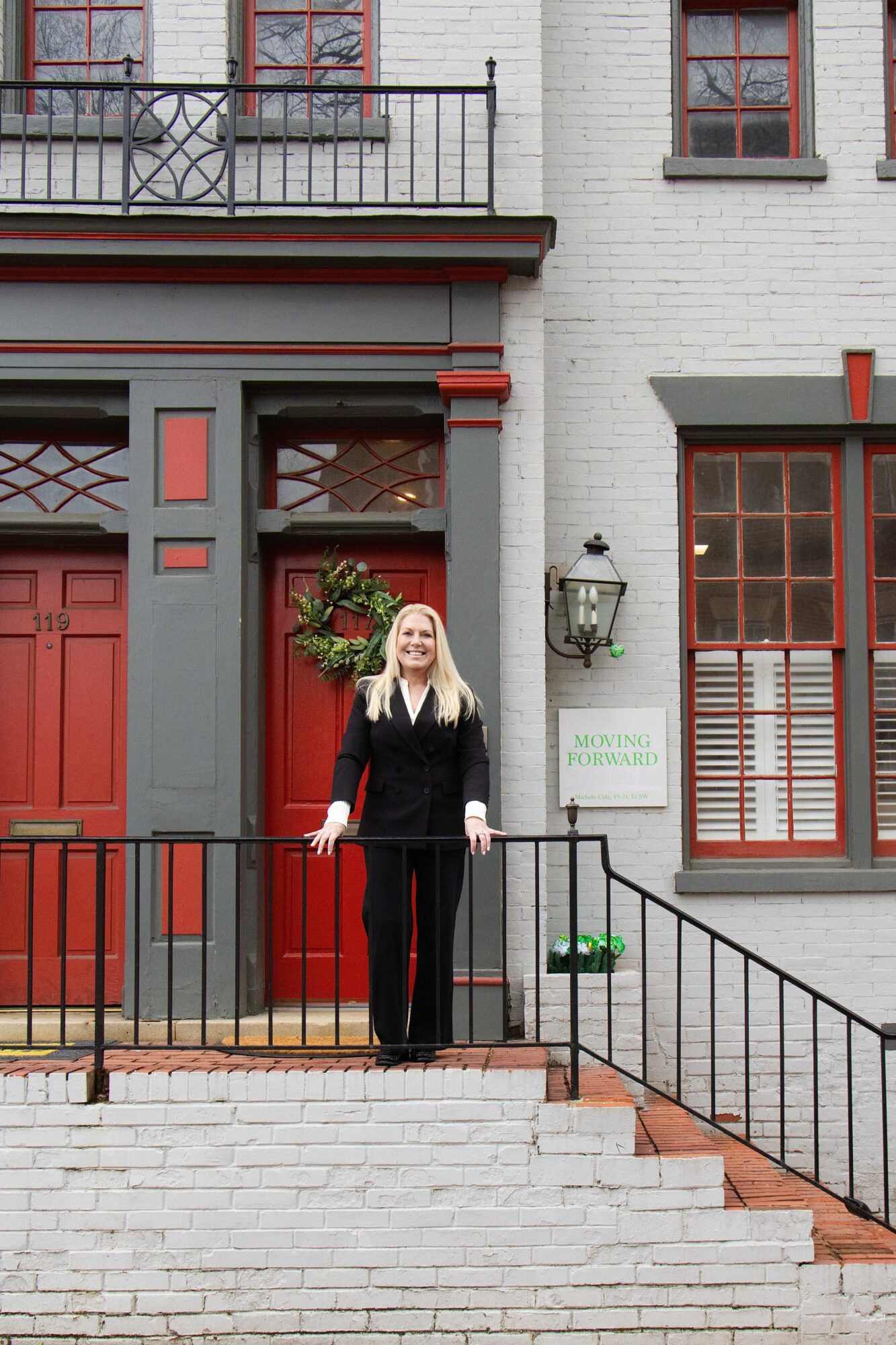 A person stands on a raised brick stoop in front of a gray and red building with windows and black railings.